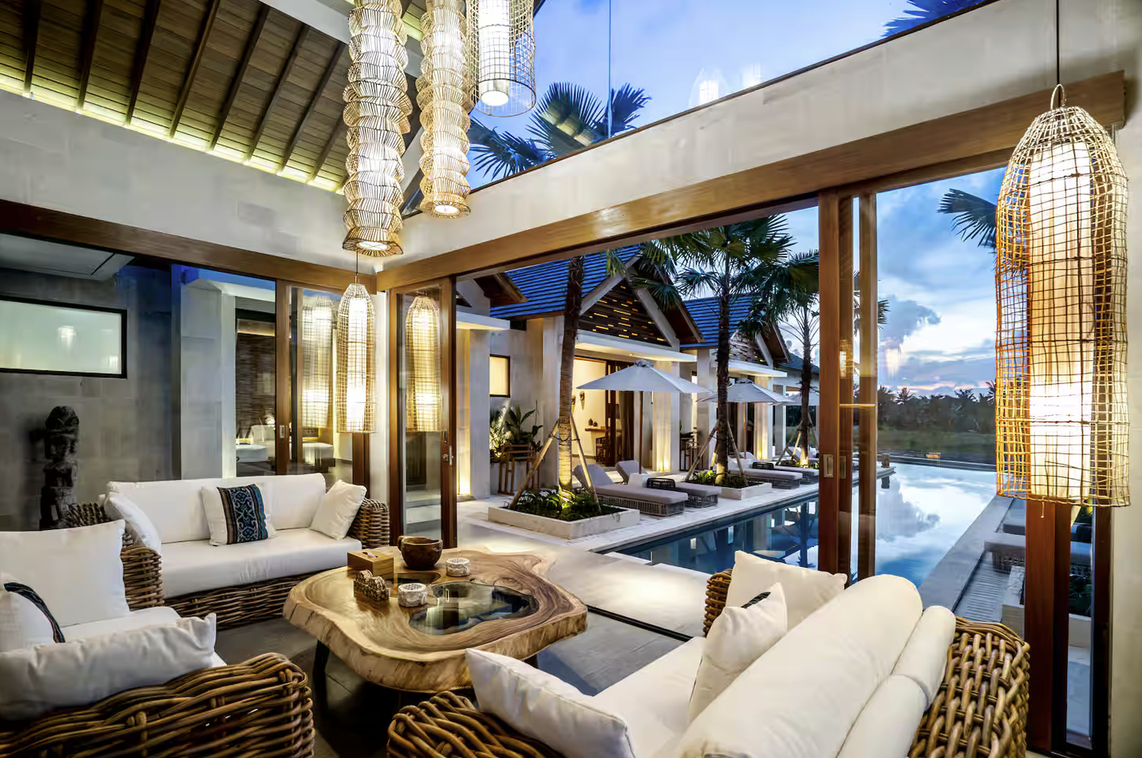 An indoor-outdoor tropical lounge area with rattan furniture, decorative pendant lights, and a pool surrounded by palm trees and lounge chairs.