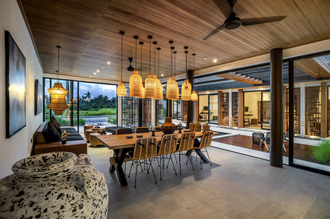 Modern dining room with a large wooden table, rattan chairs, hanging pendant lights, a large ceramic vase, and large sliding glass doors leading to an outdoor patio with greenery.
