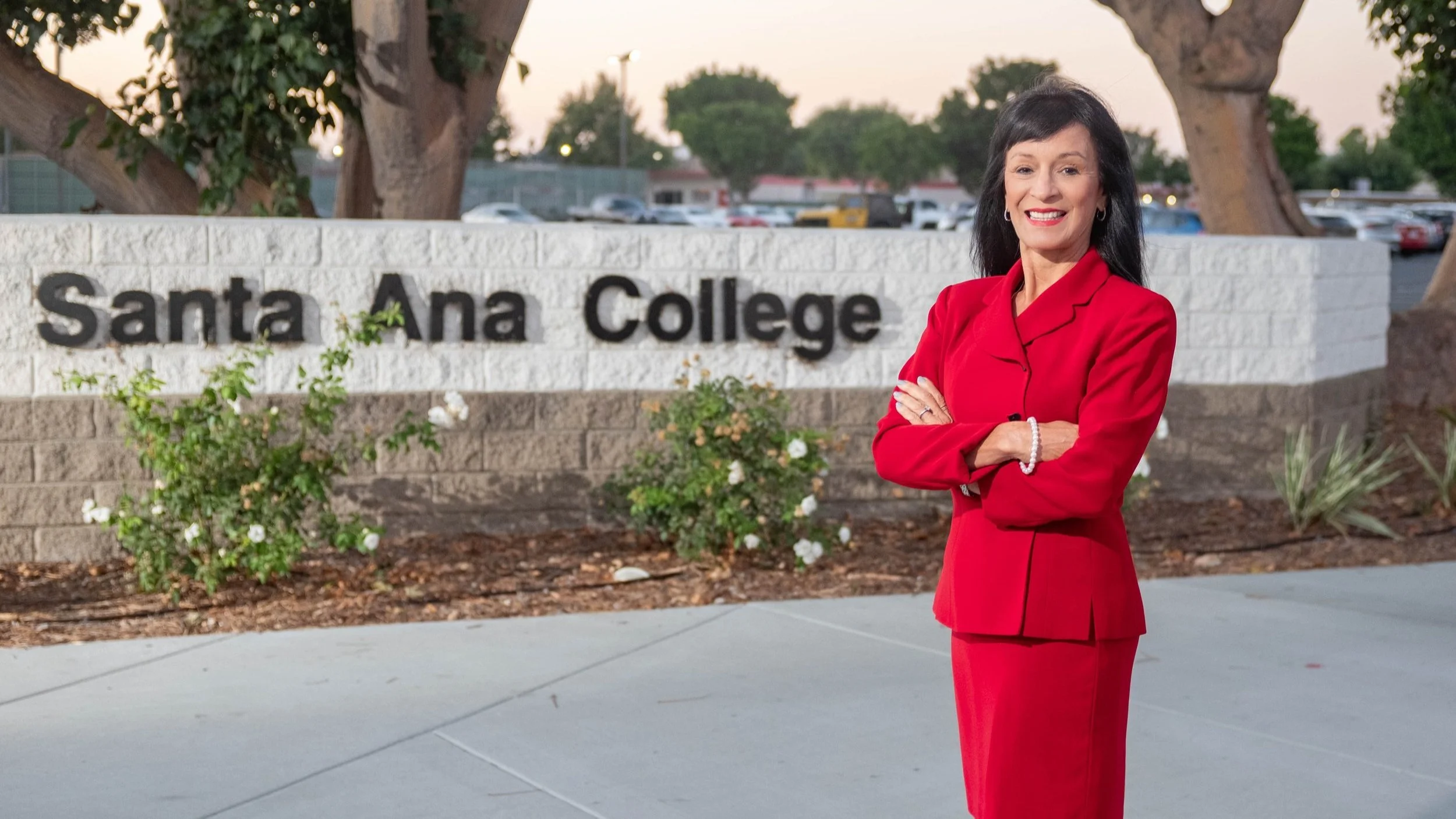 A woman in a red blazer and skirt standing outdoors in front of a sign that reads "Santa Ana College." She has dark hair, is smiling, and has her arms crossed.