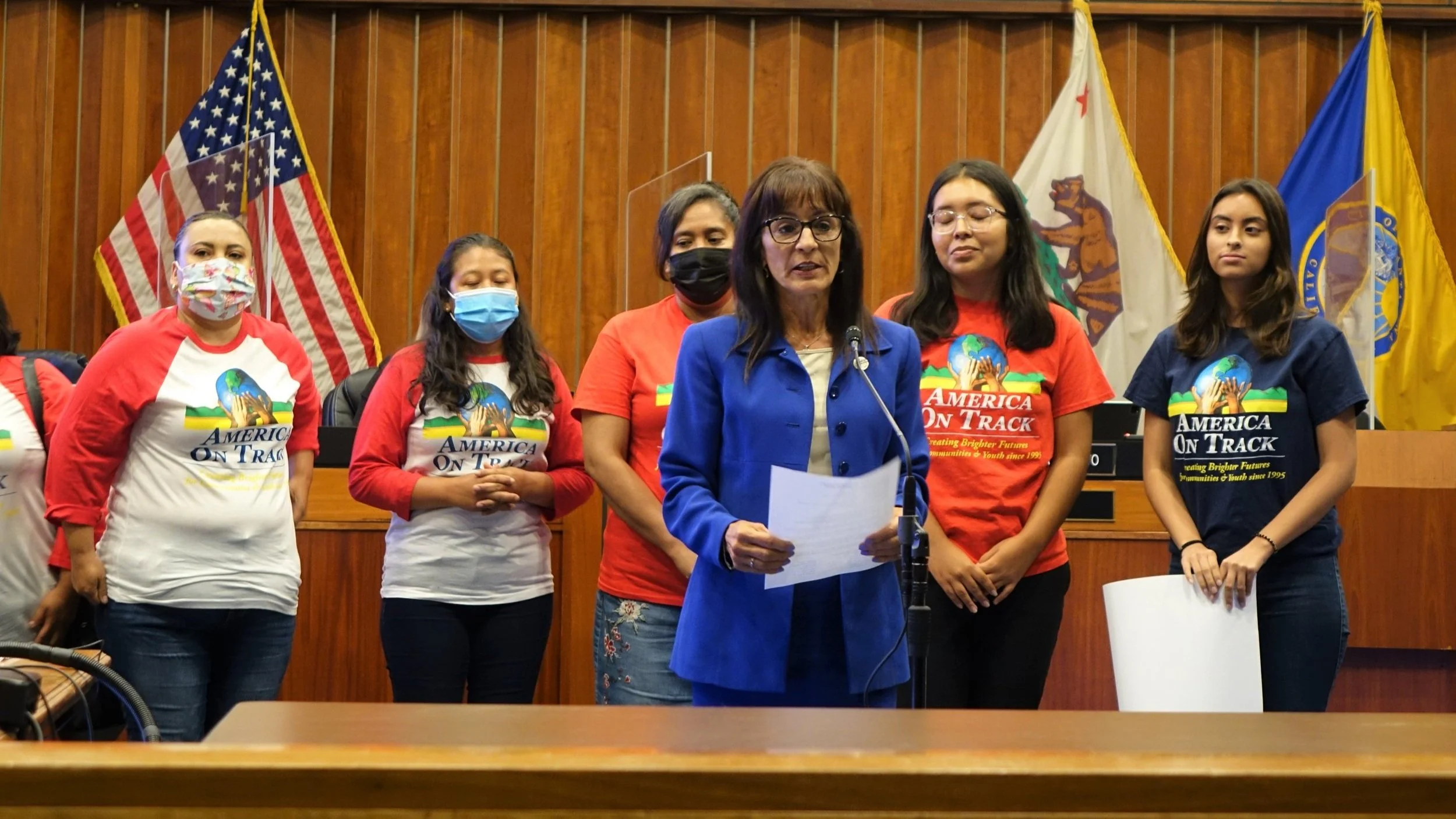Women and girls standing in a wooden-paneled room with flags in the background, some wearing face masks and Red and White 'America on Track' T-shirts, while a woman in a blue jacket speaks at a microphone.