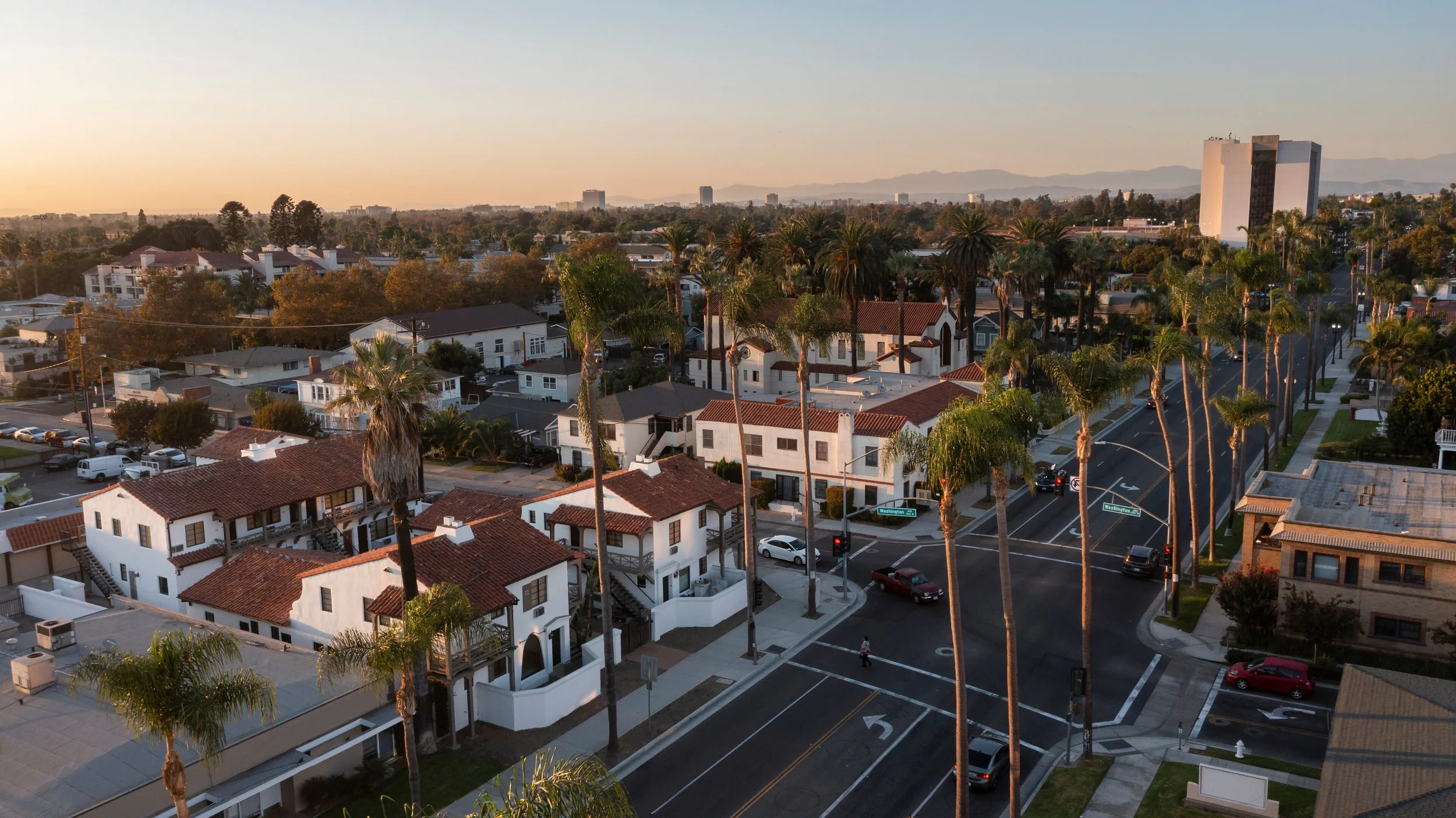 Wide view of a city neighborhood at sunset with white houses, palm trees, and a mountains in the background.