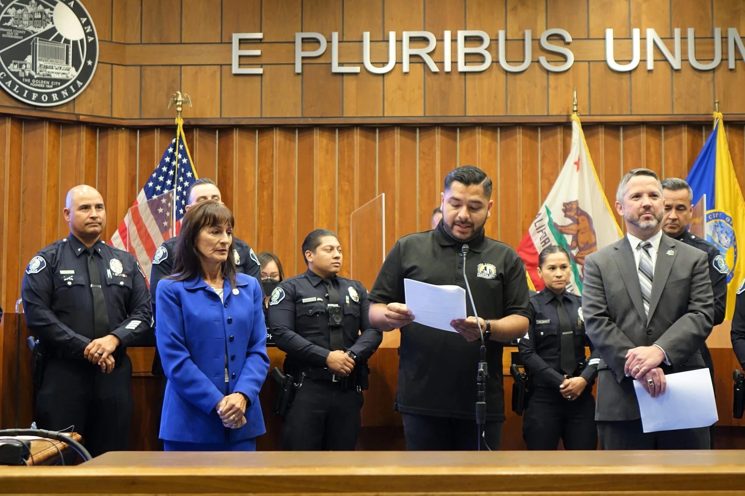 A group of police officers and officials standing in a courtroom or government chamber, with the sign 'E Pluribus Unum' and flags behind them.