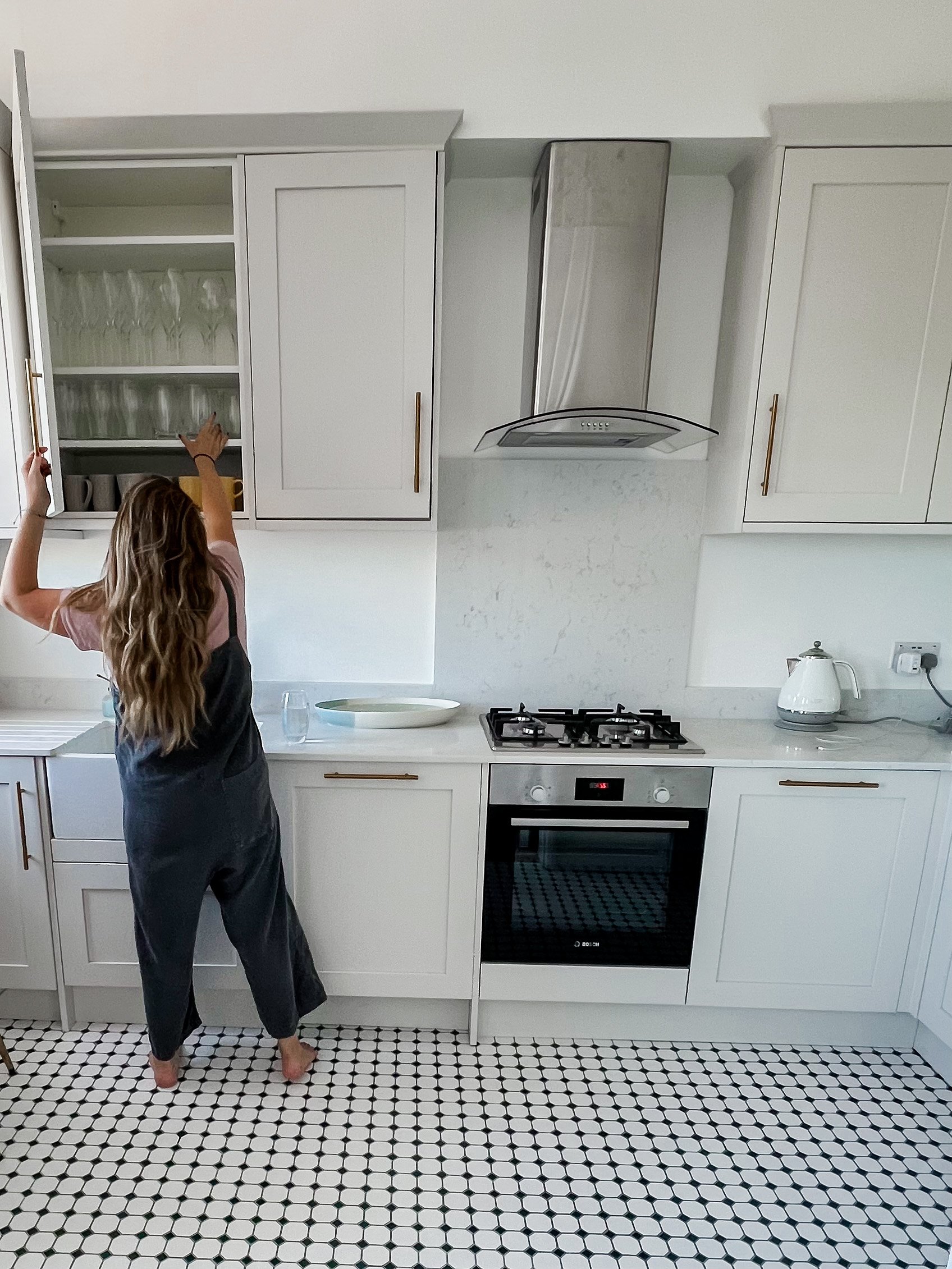 A woman with long wavy hair reaching into an open kitchen cabinet above the counter, which contains glasses and cups, in a modern white kitchen with black and white tiled floor.