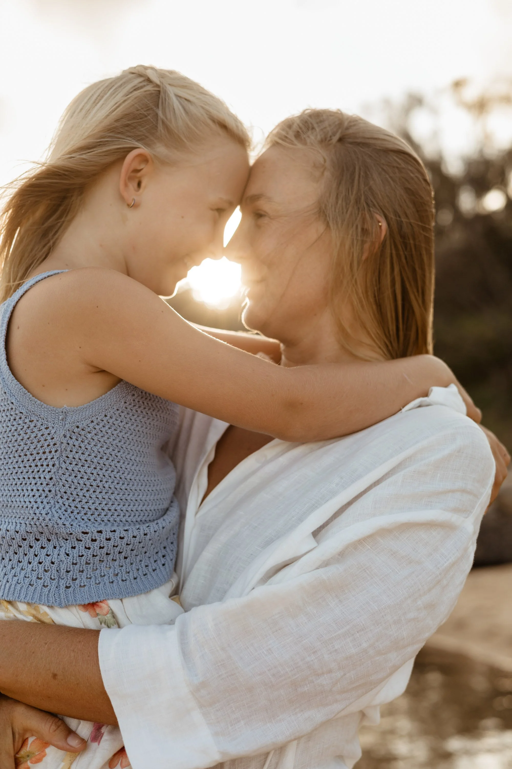 A close-up of a mum and daughter, with the sun peeking through the middle of their faces. The woman is wearing a white shirt and the daughter is wearing a blue knit singlet top. Photo by Sunshine Coast Photographer.