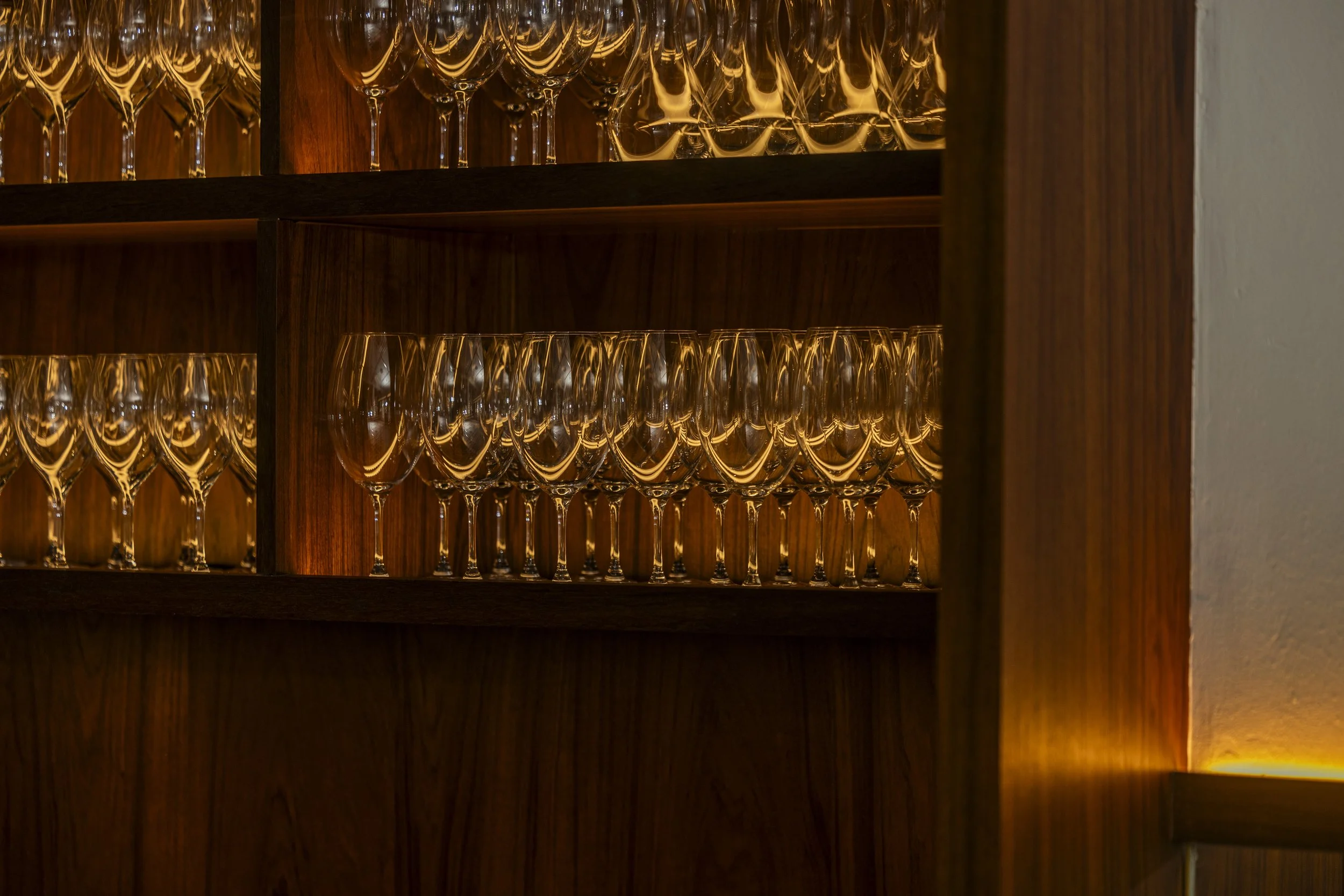 Multiple champagne flutes and wine glasses stored on wooden shelves in a cabinet at Chip Bee Bistro, with some glasses tinted with a golden hue from interior lighting.