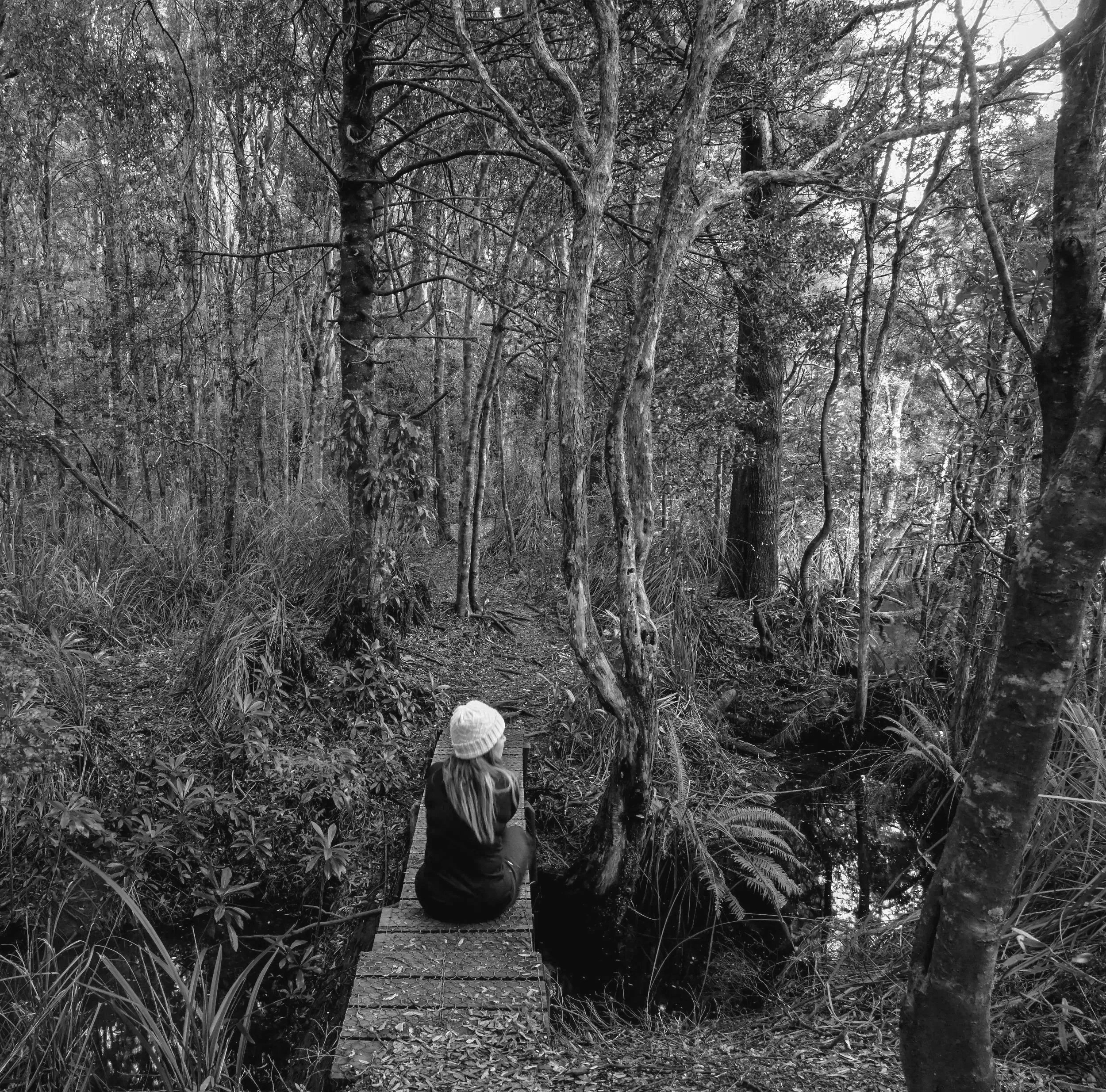 A woman with long hair wearing a beanie hat and dark clothing sits on a small bridge in a dense forest, facing a narrow creek surrounded by tall trees and thick foliage.