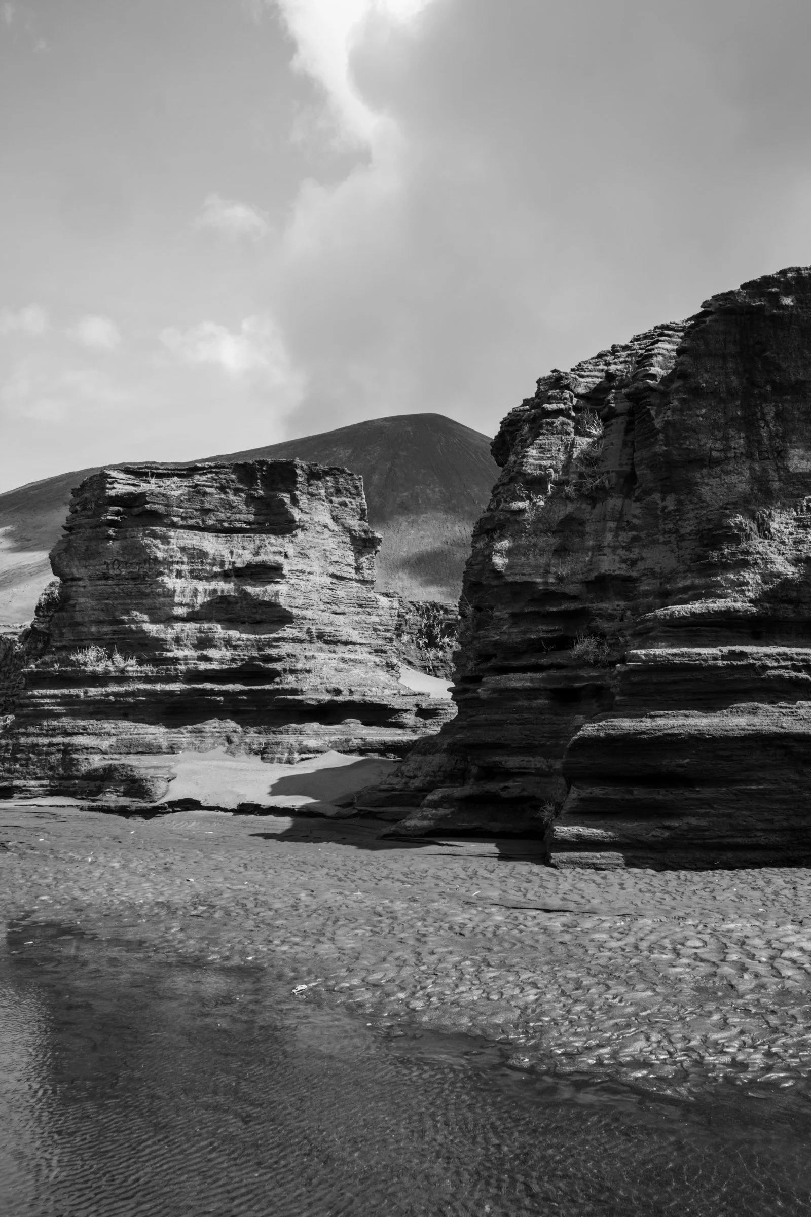 Black and white photograph of cliffs and rock formations by a body of water, with a mountain in the background and partly cloudy sky.
