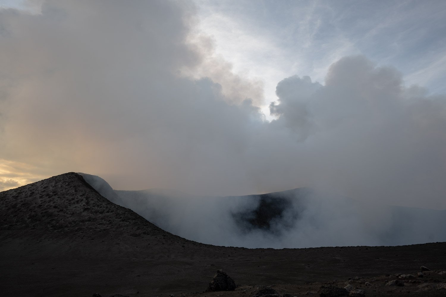 A volcanic landscape with dark, rocky terrain, erupting smoke and ash, under a cloudy sky with some light breaking through.