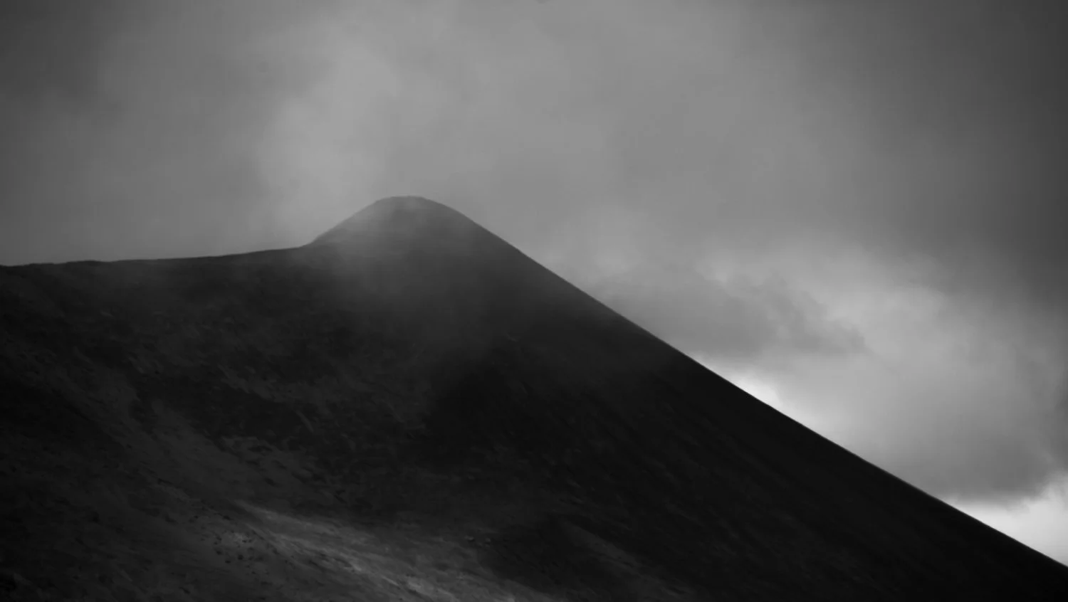 A black and white photo of a mountain with a conical peak, partially obscured by clouds and mist.