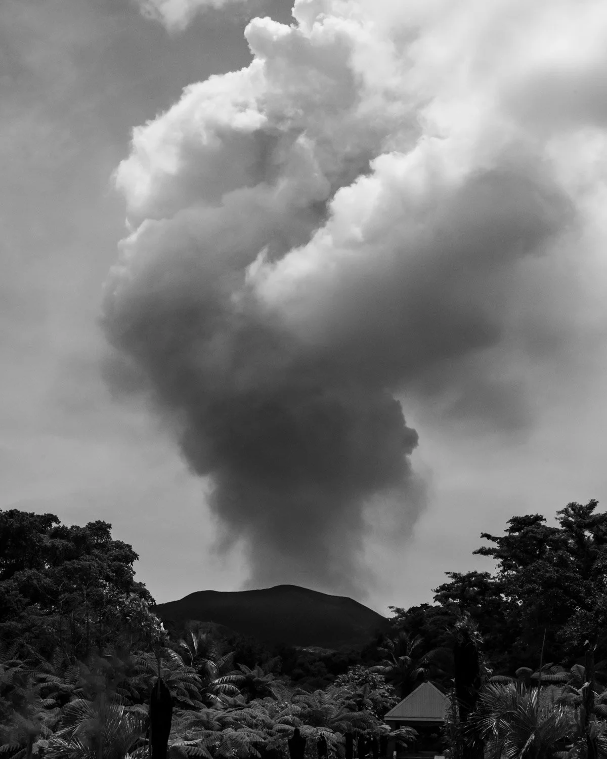 Black and white photo of a large cloud of smoke rising above a landscape with trees and hills in the foreground.