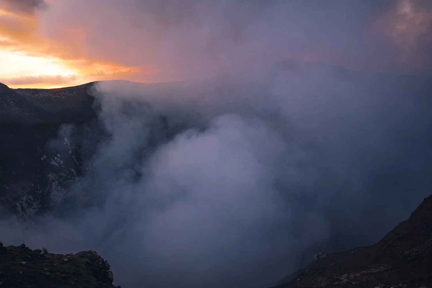 Volcanic crater with smoke and ash, with a colorful sunset sky in the background.