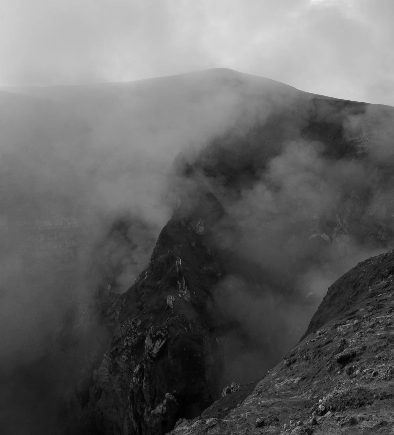 A black and white photo of a mountain with steep rocky sides, shrouded in mist or clouds, creating a dramatic and mysterious atmosphere.