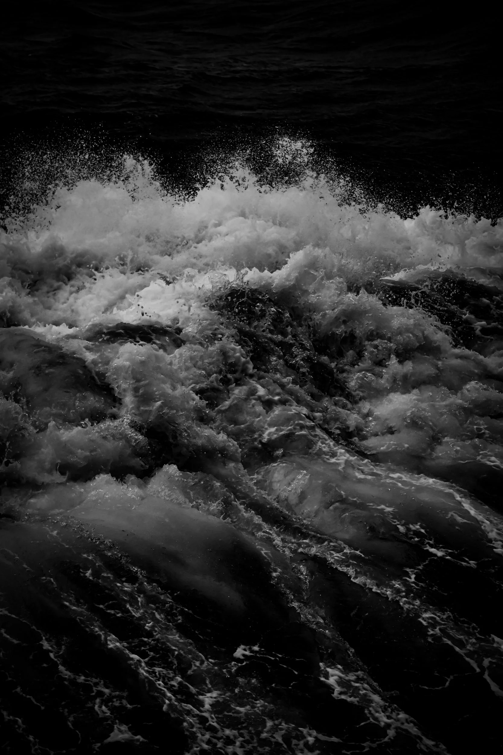 Black and white photo of ocean waves crashing against rocks, with foam and spray in the foreground and dark water in the background.