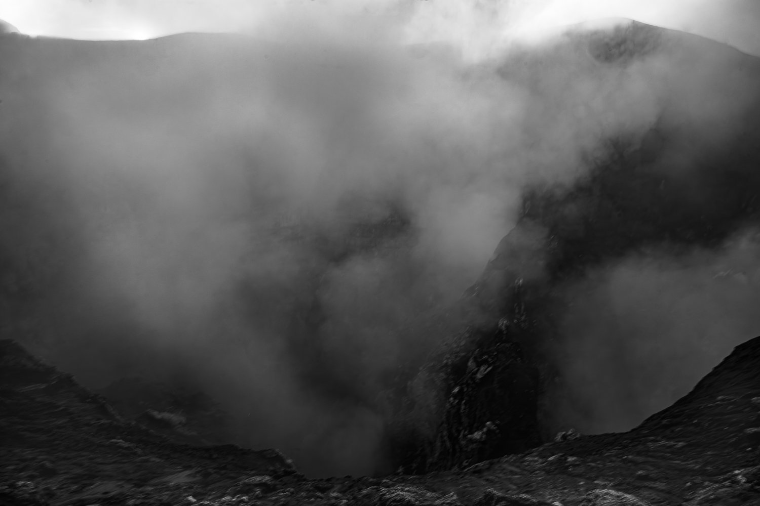 Black and white photograph of a volcanic eruption with ash and smoke billowing into the sky, dark volcanic rocks in the foreground, and a mountain peak in the background.