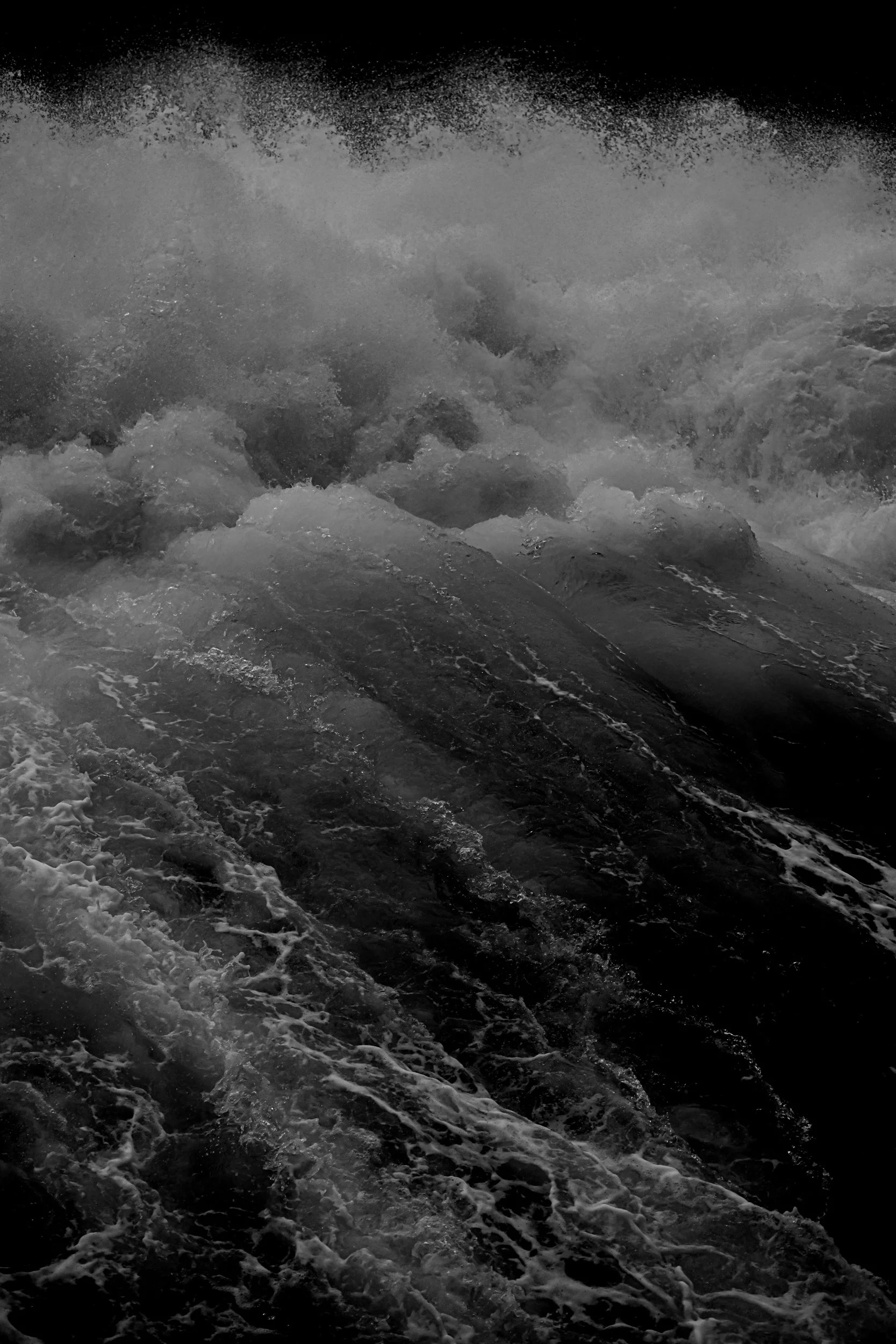Black and white photo of turbulent ocean waves crashing, foam and spray visible at the surface.