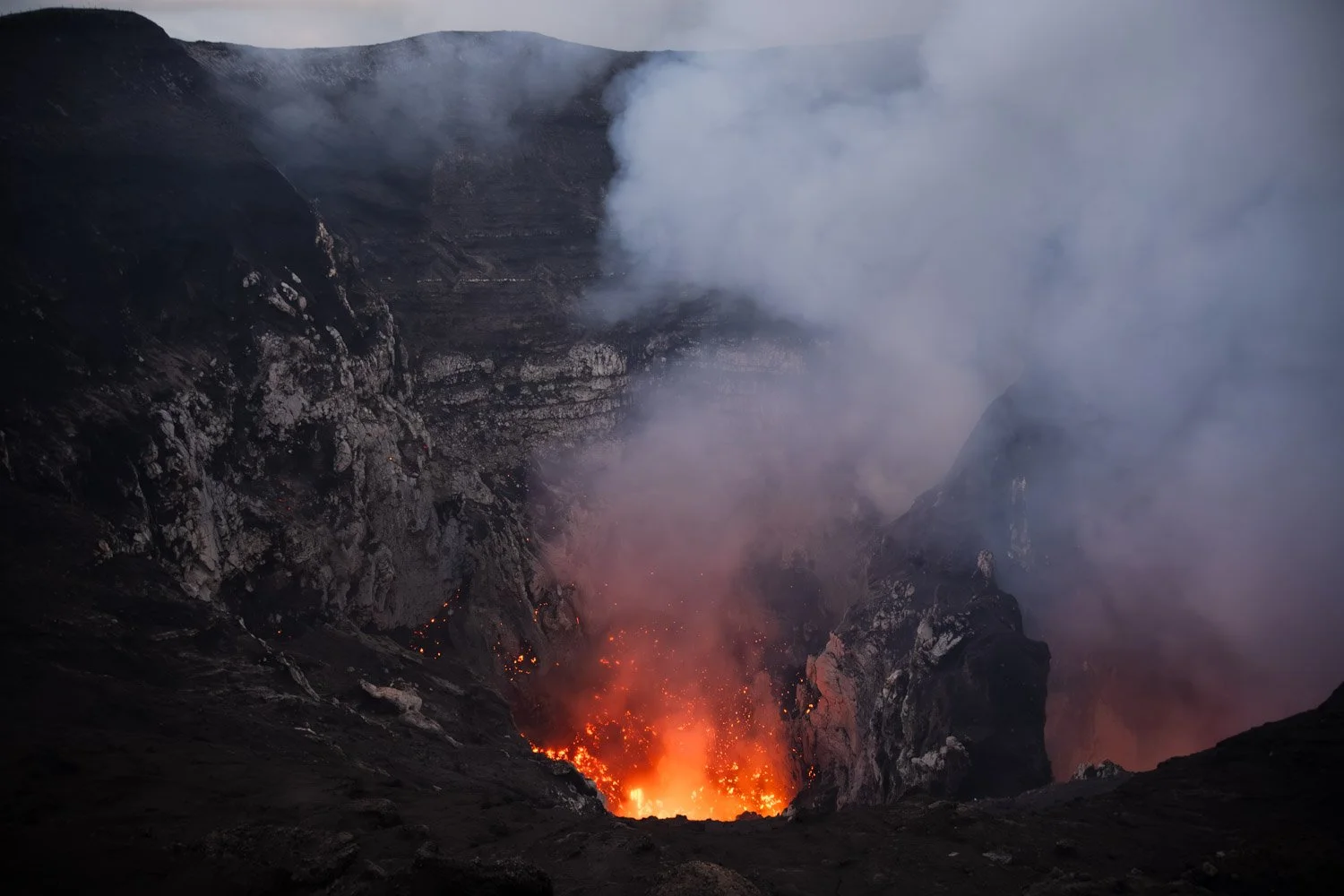 Volcano erupting with flowing lava, smoke, and ash in a dark, mountainous landscape.