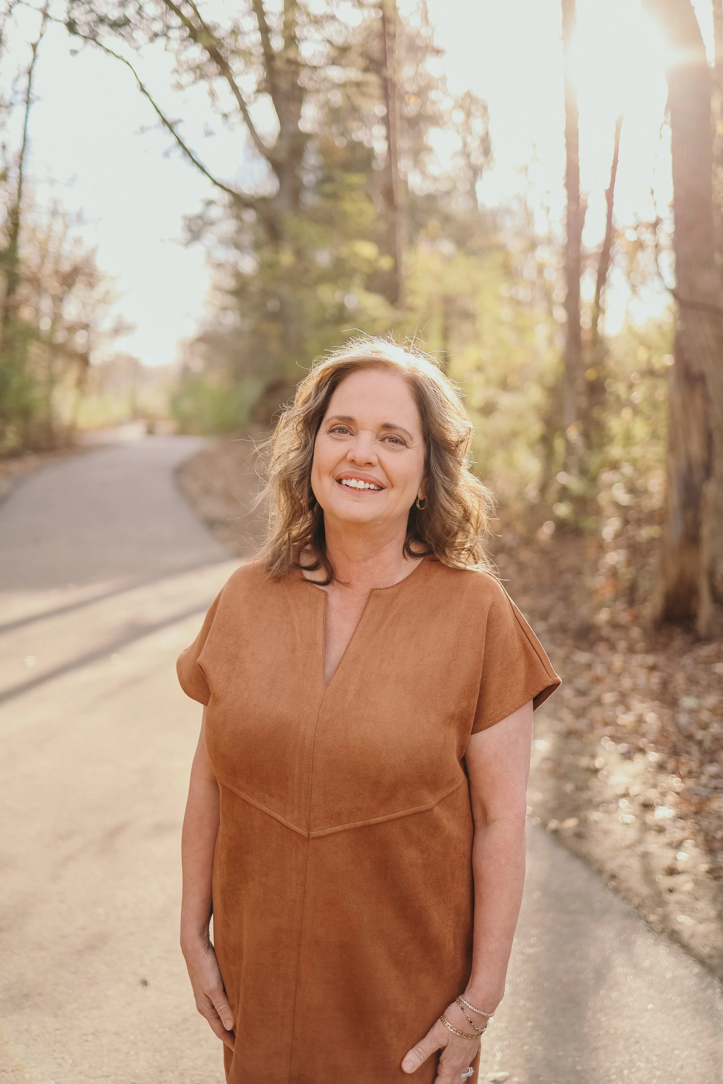 Portrait of a smiling woman with blonde hair wearing pearl earrings and a pearl necklace, outdoors with greenery in the background.