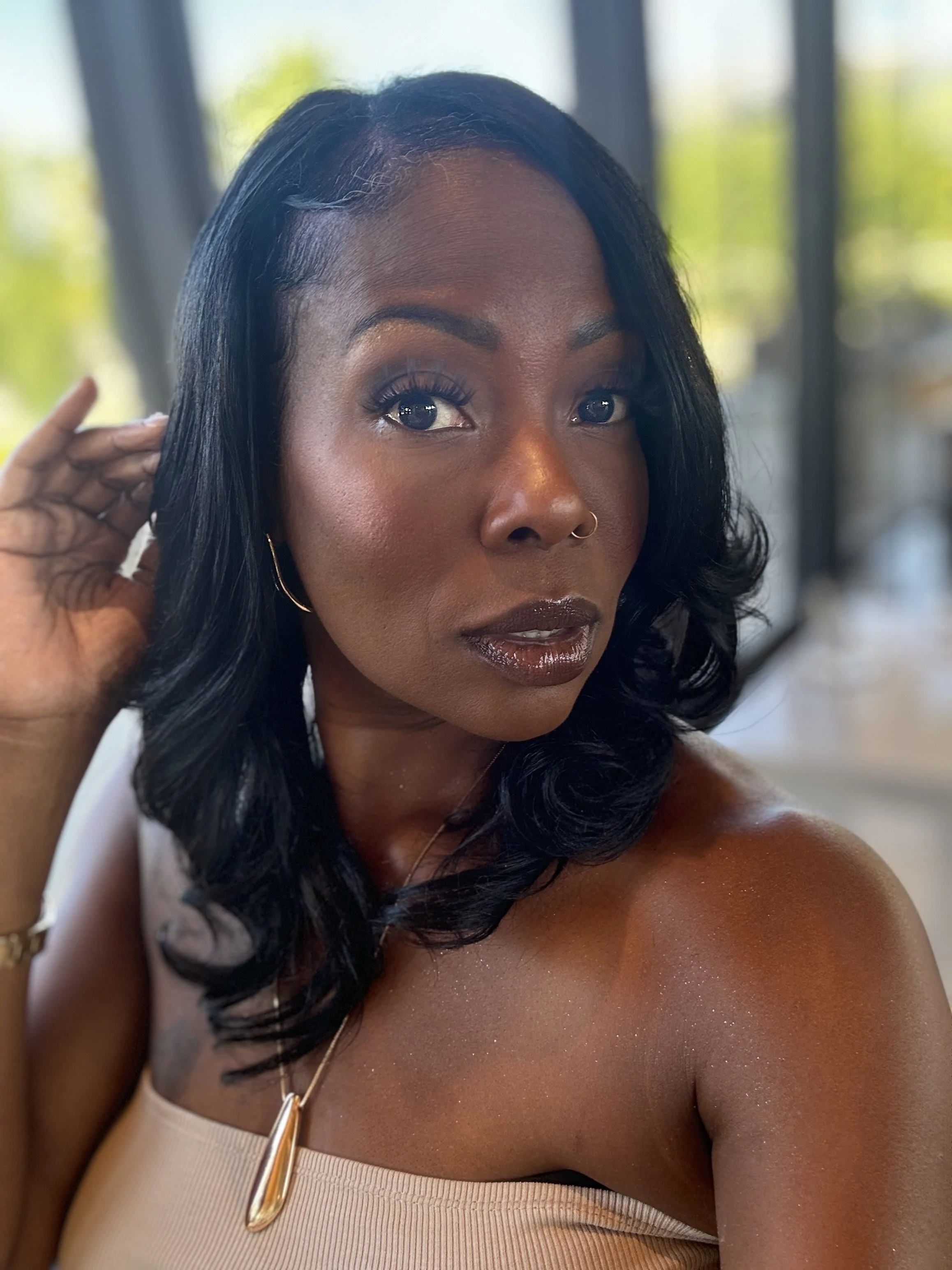 A woman with dark hair styled in loose waves, wearing makeup and gold jewelry, posing indoors near large windows with greenery in the background.