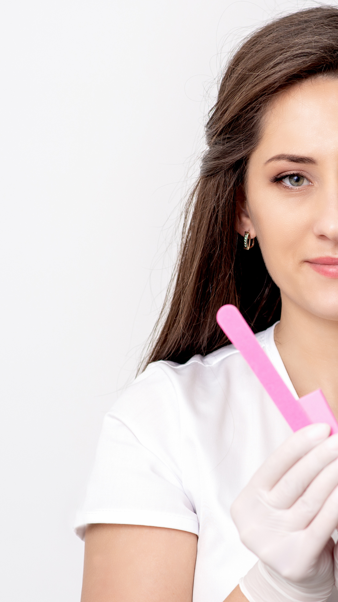 A woman with long brown hair holds a pink nail file and smiles slightly, wearing a white shirt, against a plain white background.