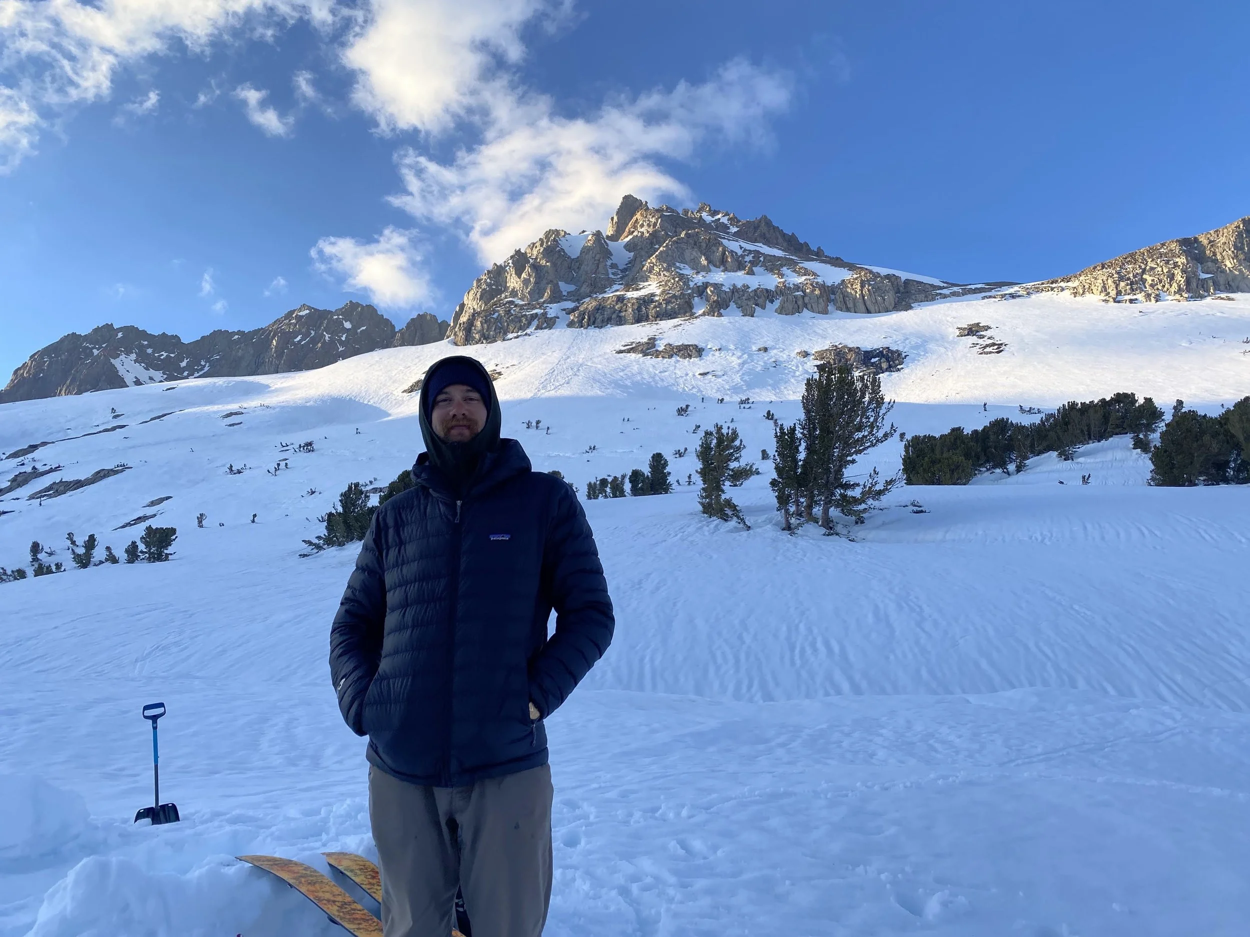 A man wearing a black jacket and standing in a snowy landscape with mountain peaks and trees in the background. Snowshoes are on the snow beside him.