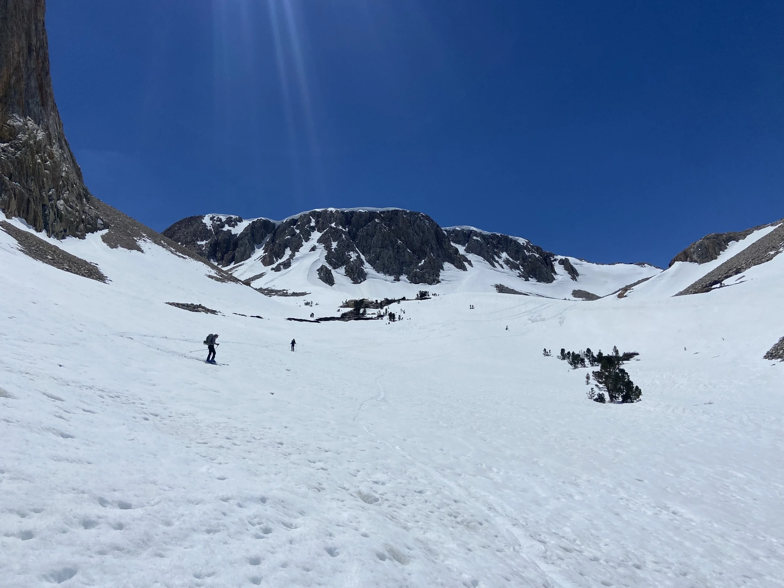 Snow-covered mountain landscape with three hikers trekking across the snow, on a clear day with a bright blue sky and rocky peaks in the background.
