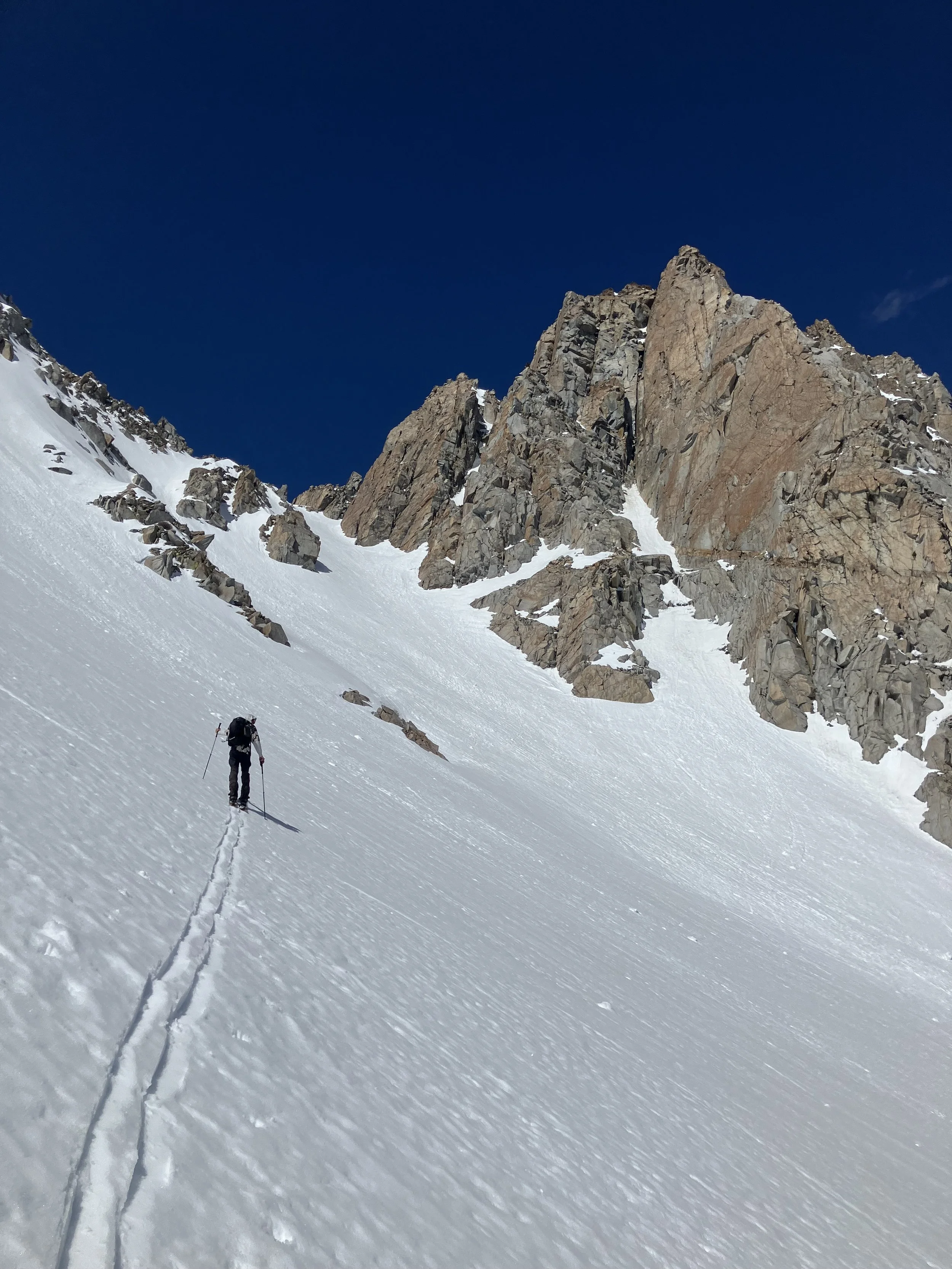 Hiker in snow-covered mountain landscape with steep slopes, rocky outcroppings, and clear blue sky.