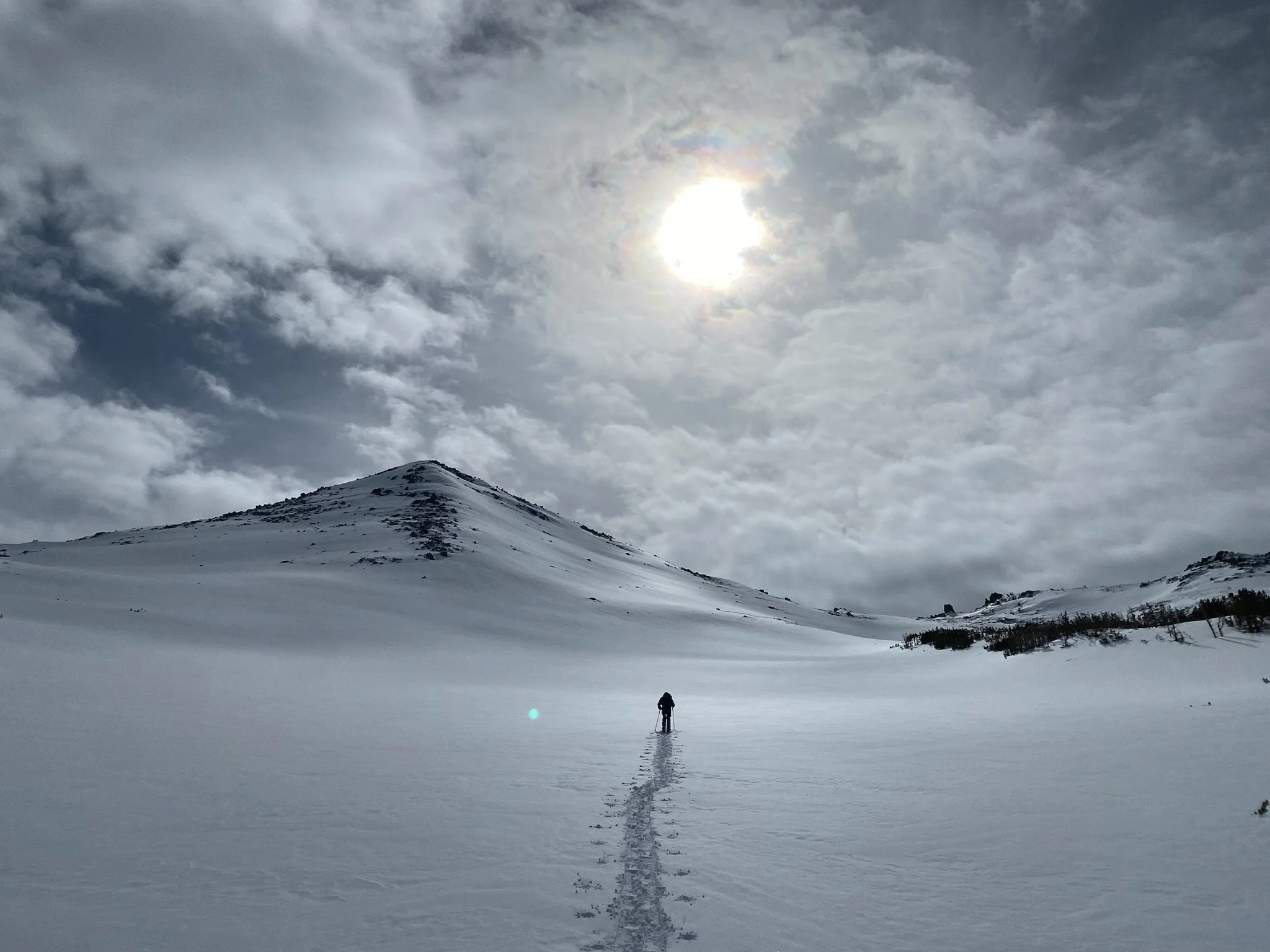 A person hiking alone in a snow-covered valley with mountain slopes, under a cloudy sky with the sun shining through.