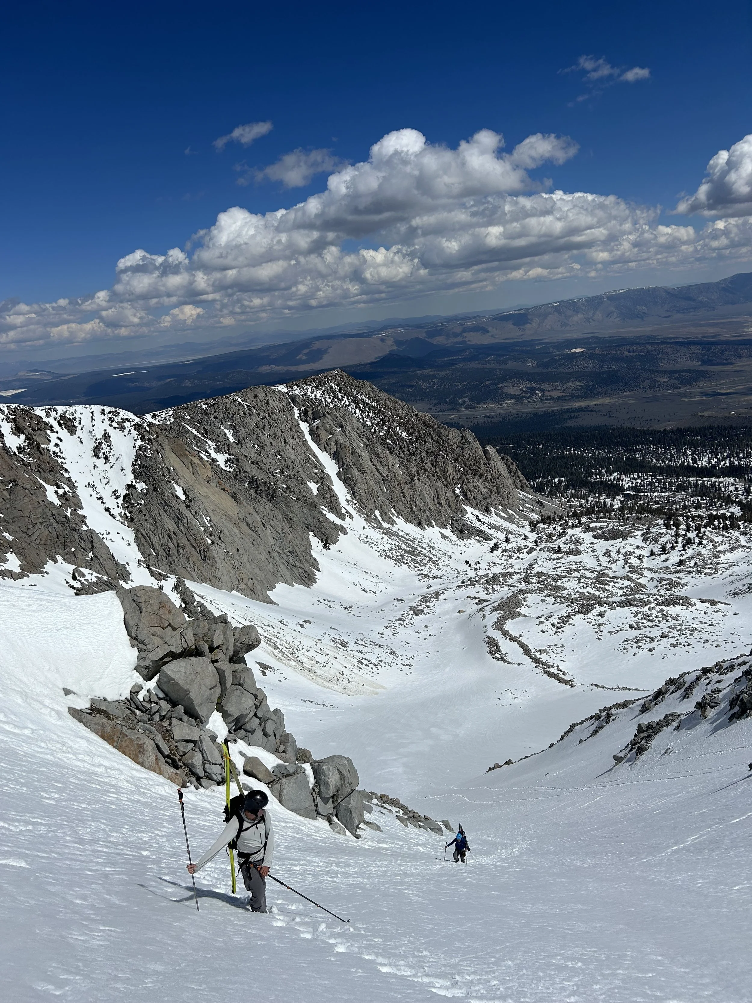 Two hikers with backpacks and trekking poles ascending a snow-covered mountain slope with rocky outcroppings, overlooking a valley and distant mountains with a partly cloudy sky.