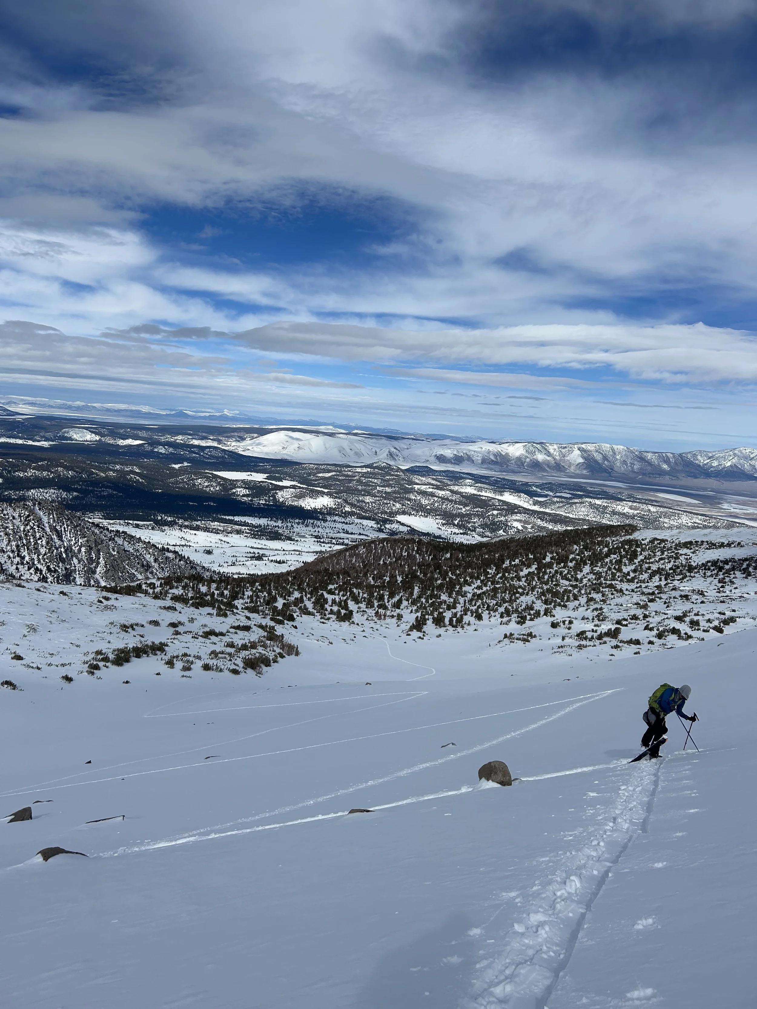 A person skiing in a snowy mountain landscape under a blue sky with scattered clouds, with distant snow-capped mountains and a valley below.
