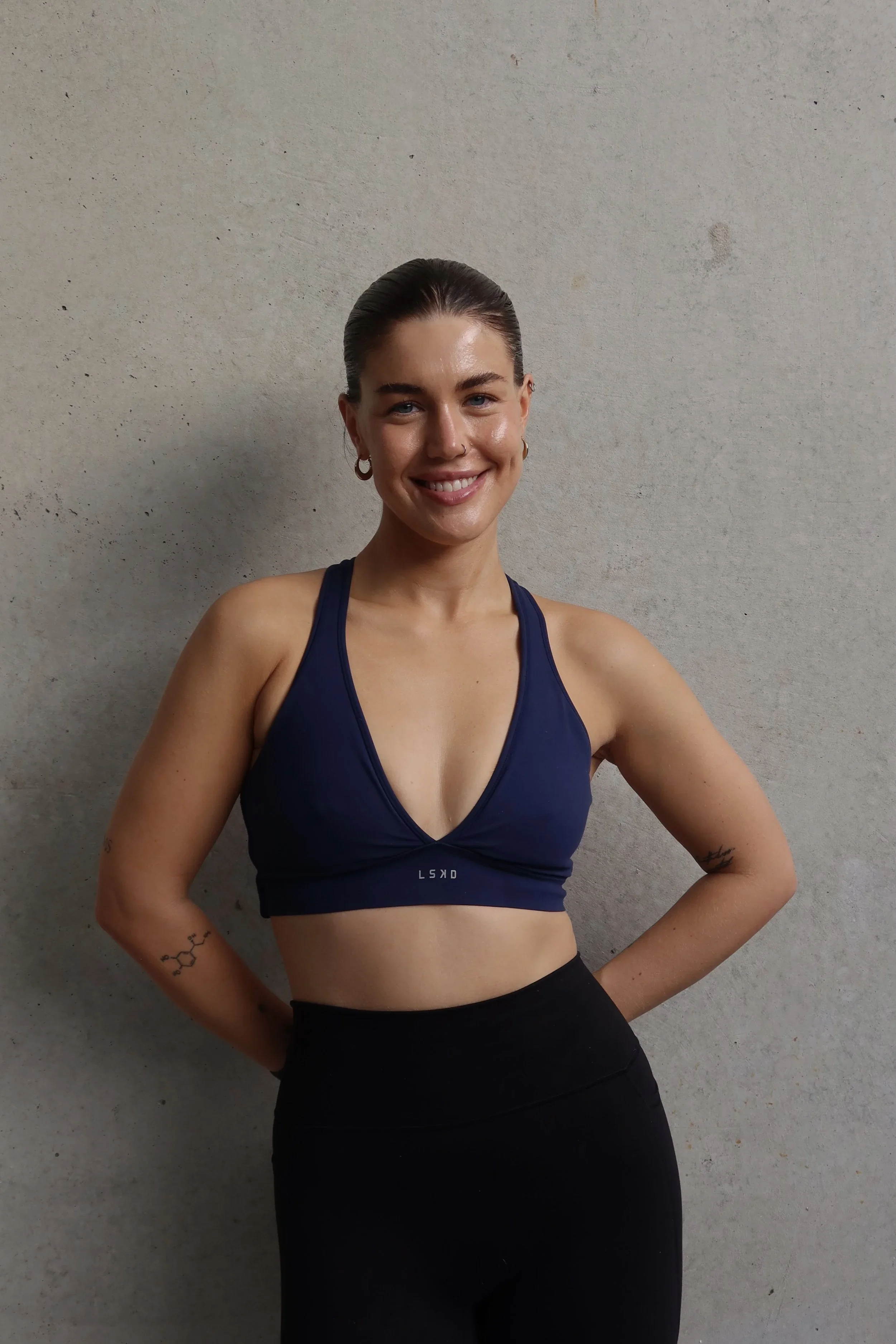 Smiling woman with dark hair in a slicked-back style, wearing a navy sports bra and black leggings, standing against a plain gray wall.