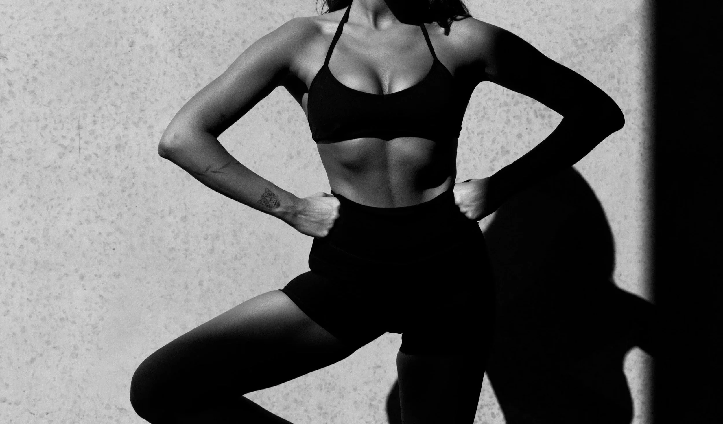 A woman is in an athletic pose, wearing a black sports bra, shorts, and tights, with shadows cast on the textured wall behind her.