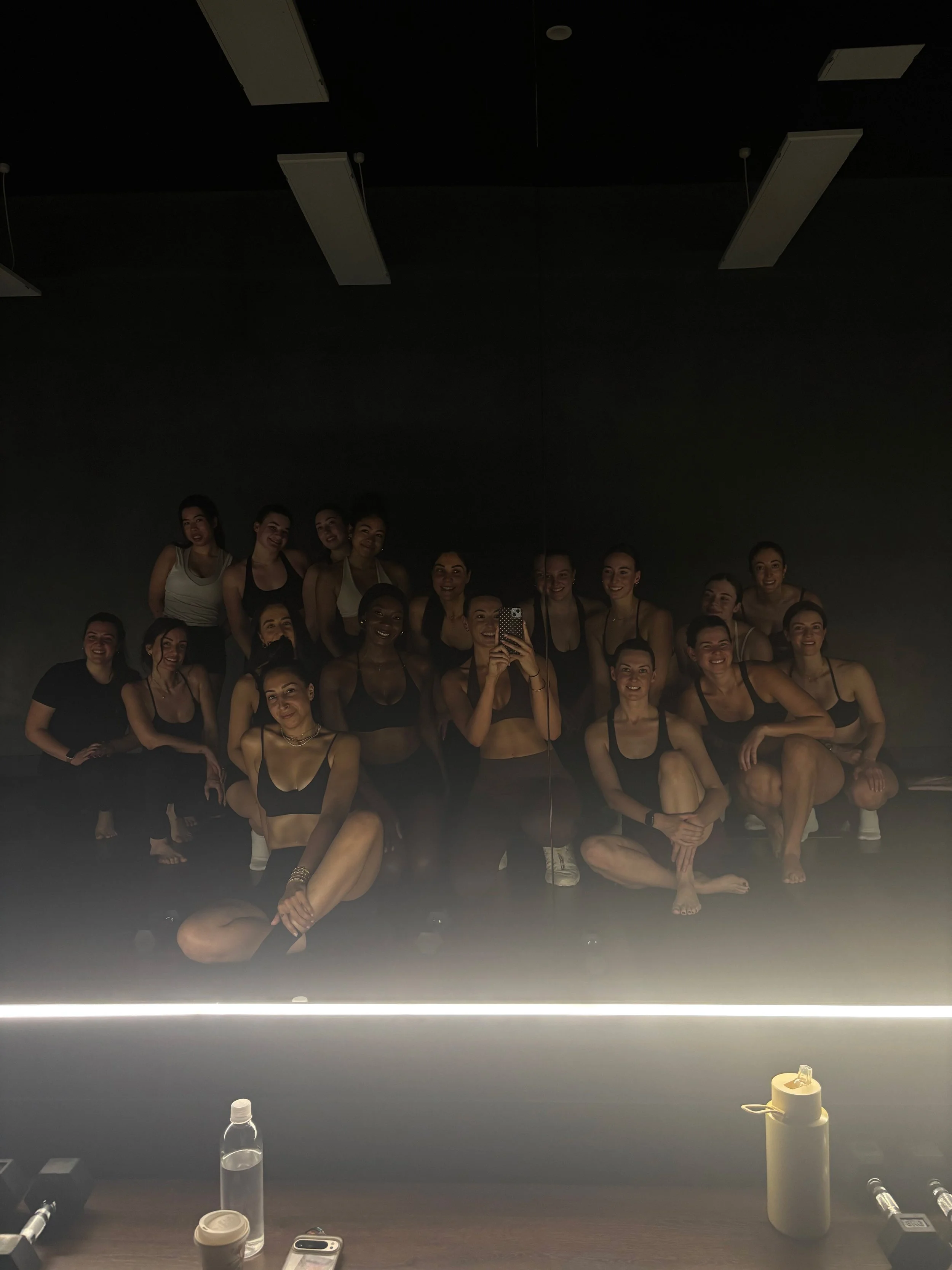 A group of women taking a mirror selfie in a dimly lit pilates studio, wearing workout or dance clothing, with water bottles, cups, and personal items on a table in front.