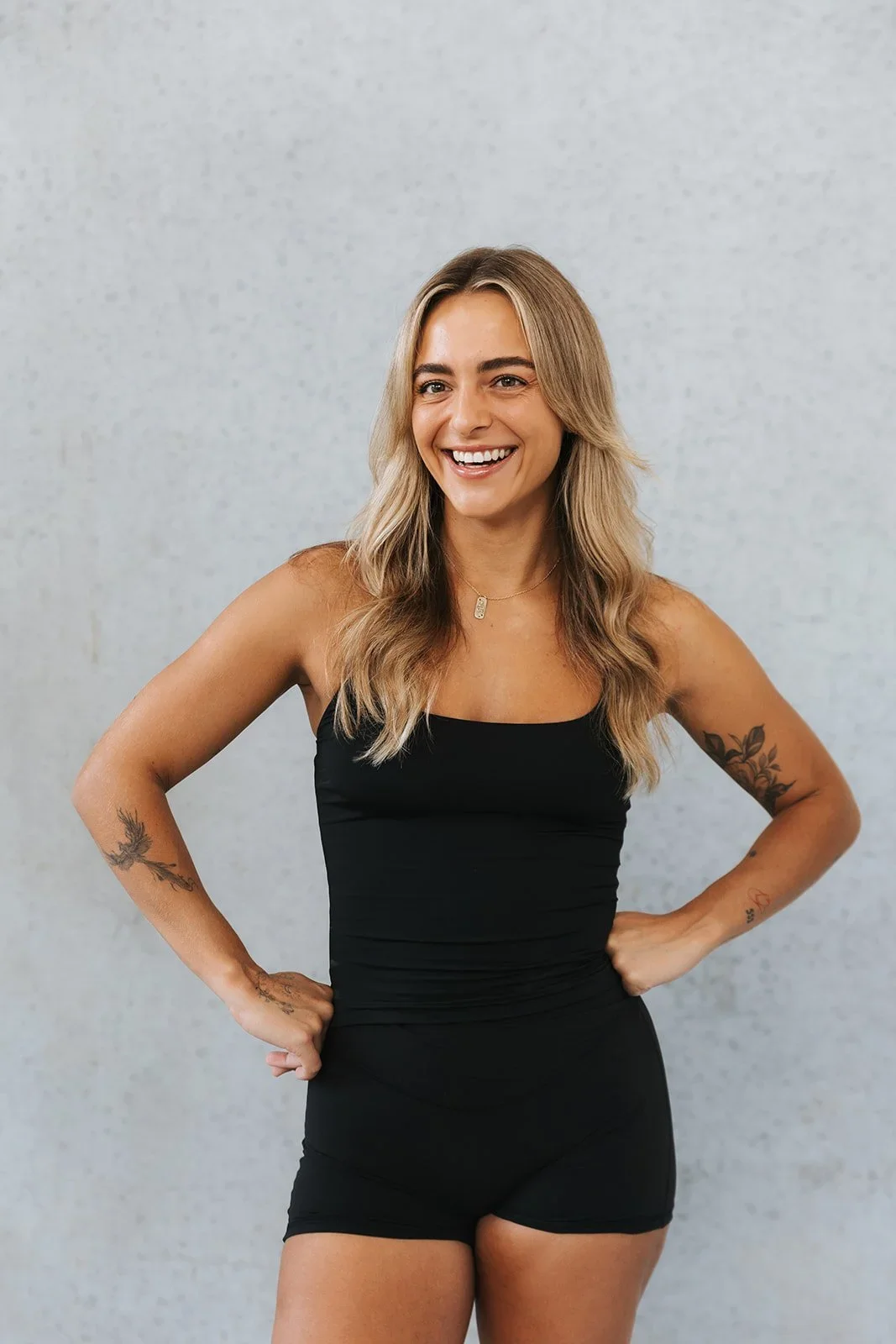 A smiling woman with wavy blond hair and tattoos on her arms, wearing a black tank top and matching shorts, stands with hands on hips against a plain light-colored background.
