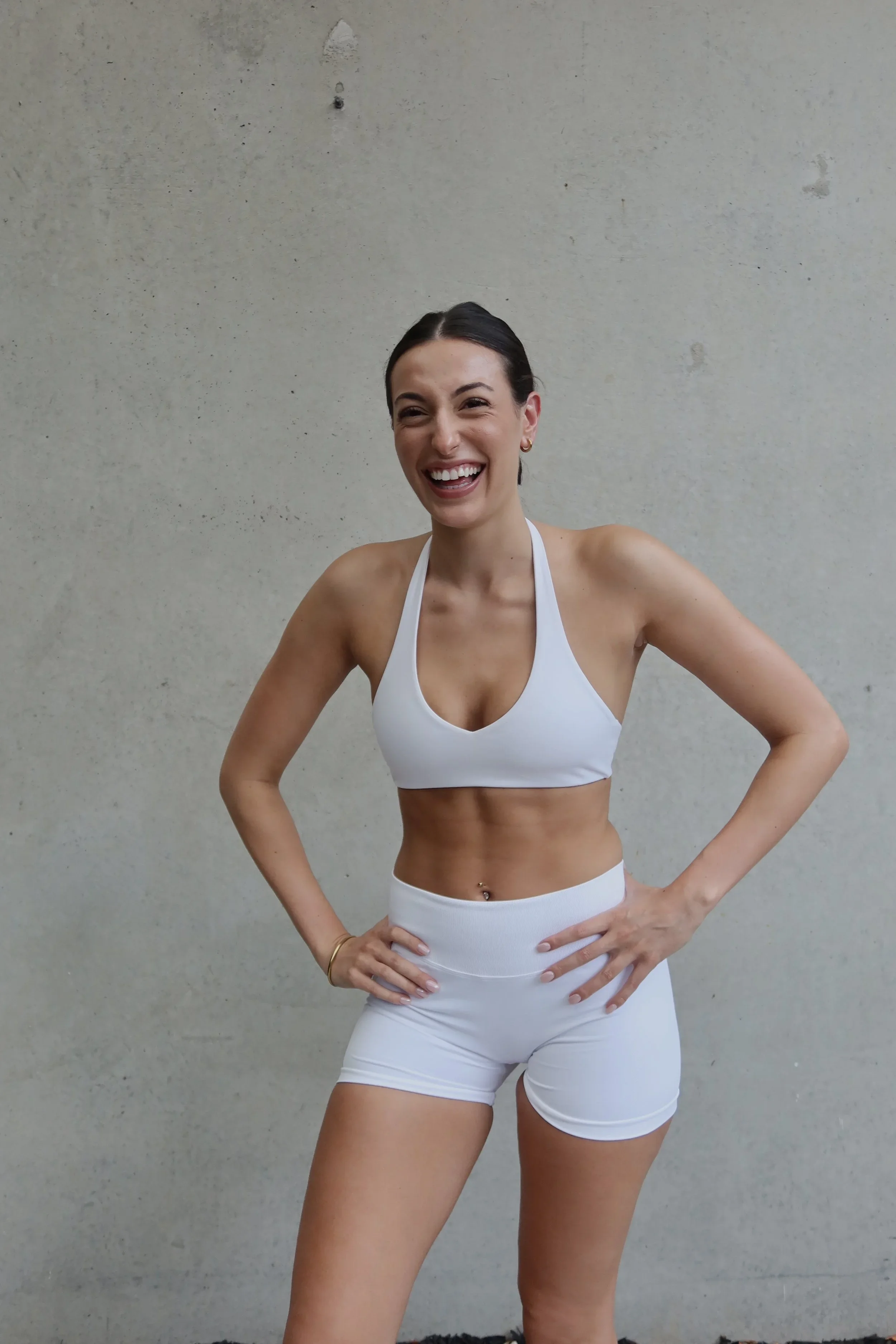 A woman with dark hair in a bun, smiling, wearing white sports bra and shorts, standing with hands on hips in front of a plain light gray wall.