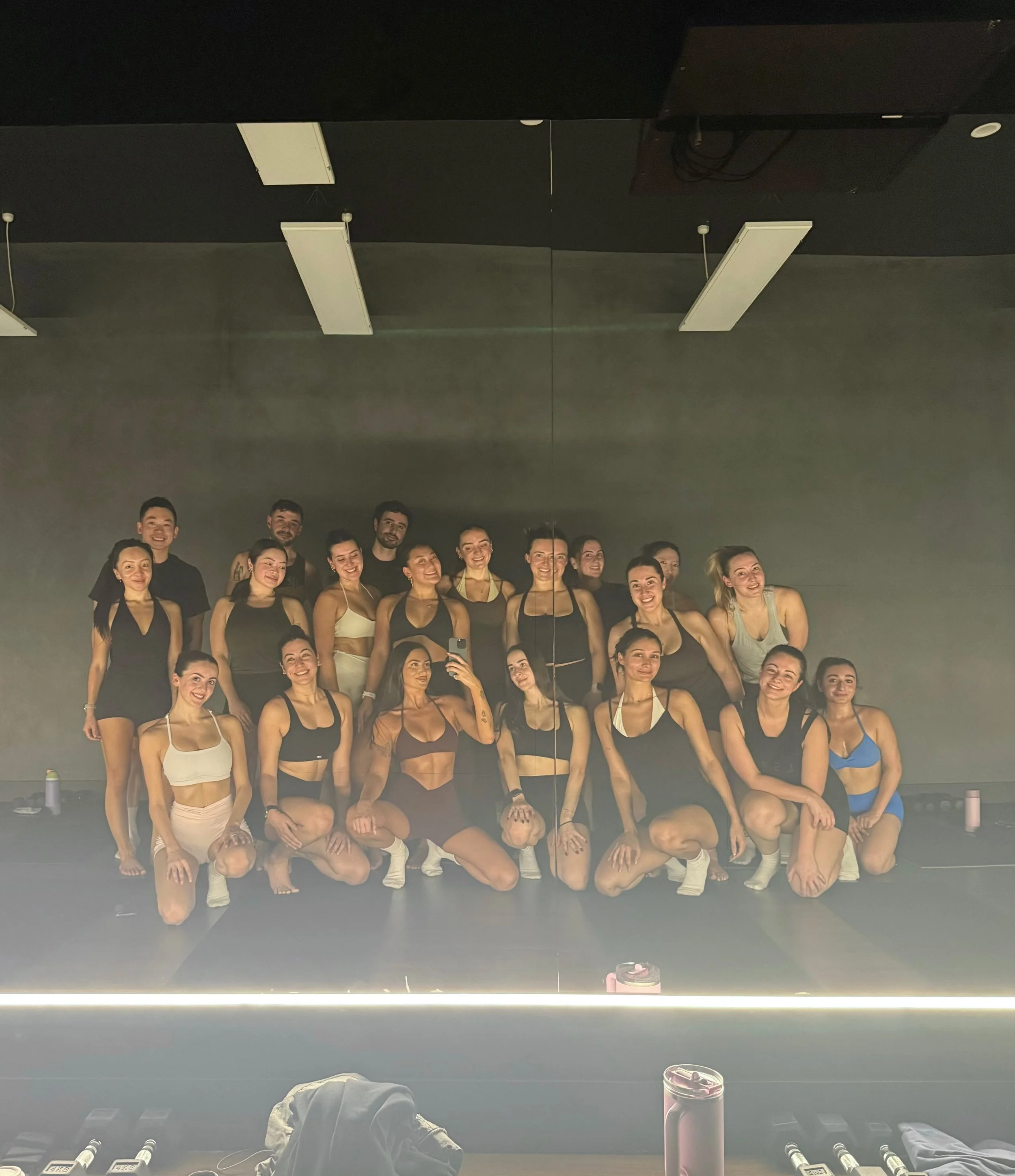 Group of 20 people in athletic wear posing for a photo in a pilates studio with a large mirror reflecting their image and ceiling lights.