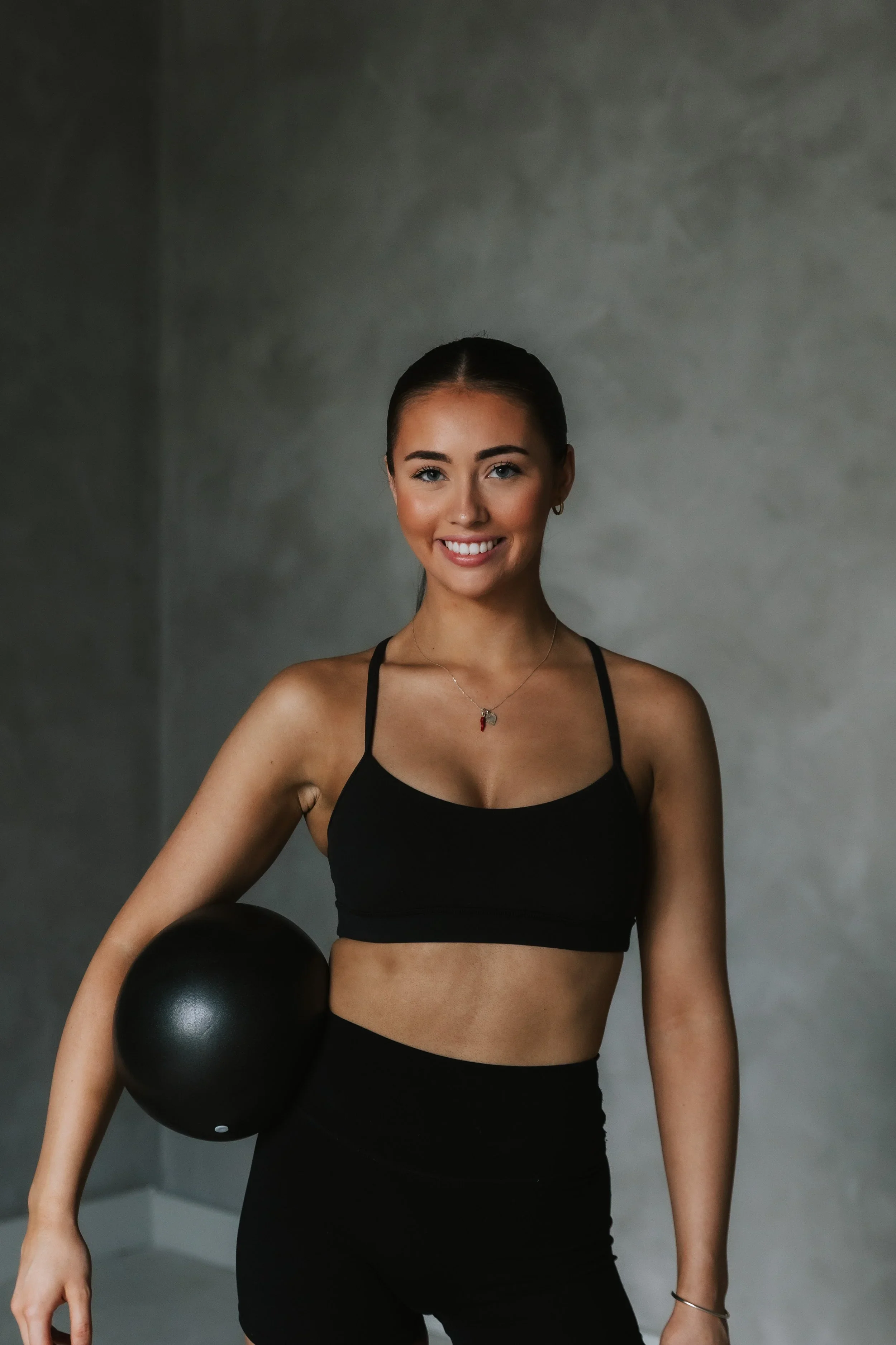 A young woman with dark hair tied back, wearing a black sports bra and black leggings, smiling, holding a black exercise ball in a minimalistic gray room.