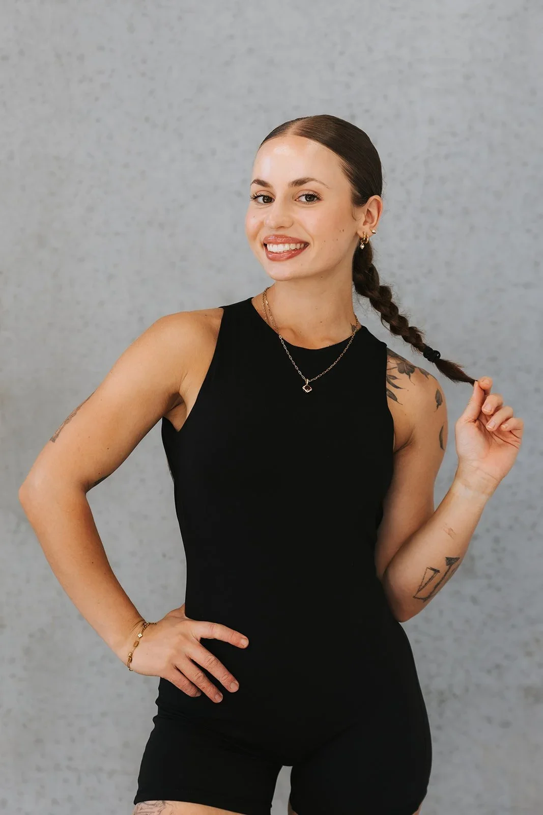 A smiling woman with brown hair in a braid wearing a black sleeveless top and black shorts, standing against a gray background.