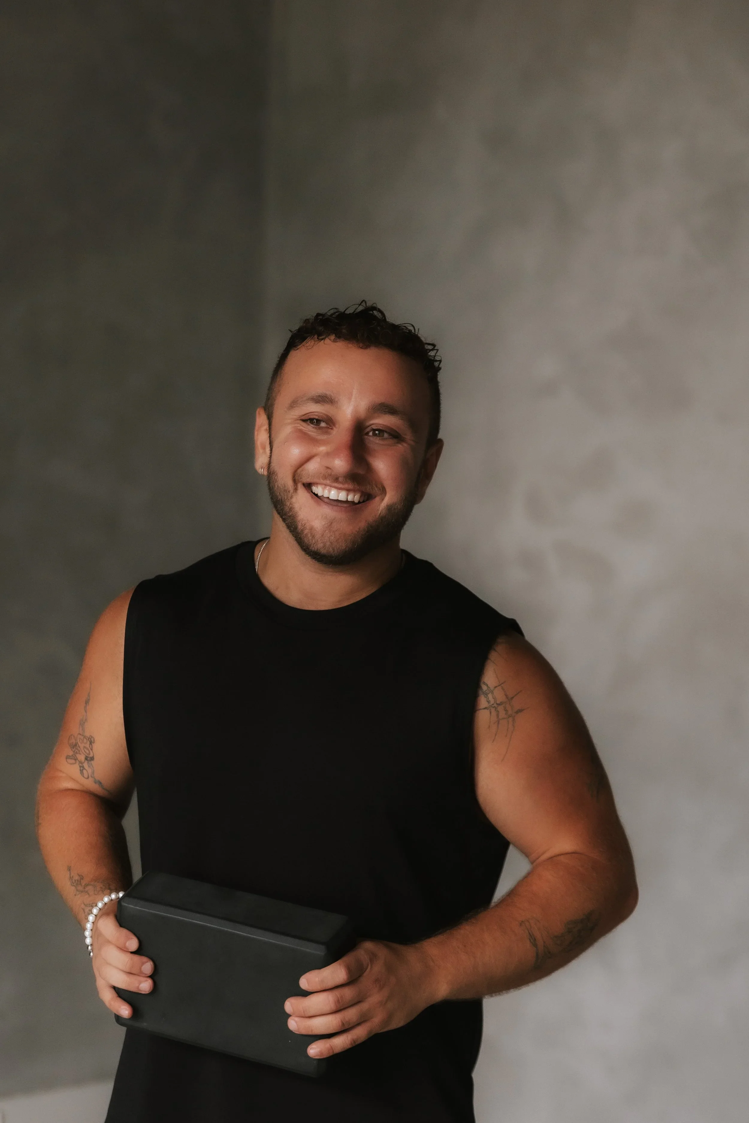 A smiling man with short curly hair, a beard, and earrings, wearing a black sleeveless shirt, holding a black box in his left hand, standing in front of a plain gray wall.