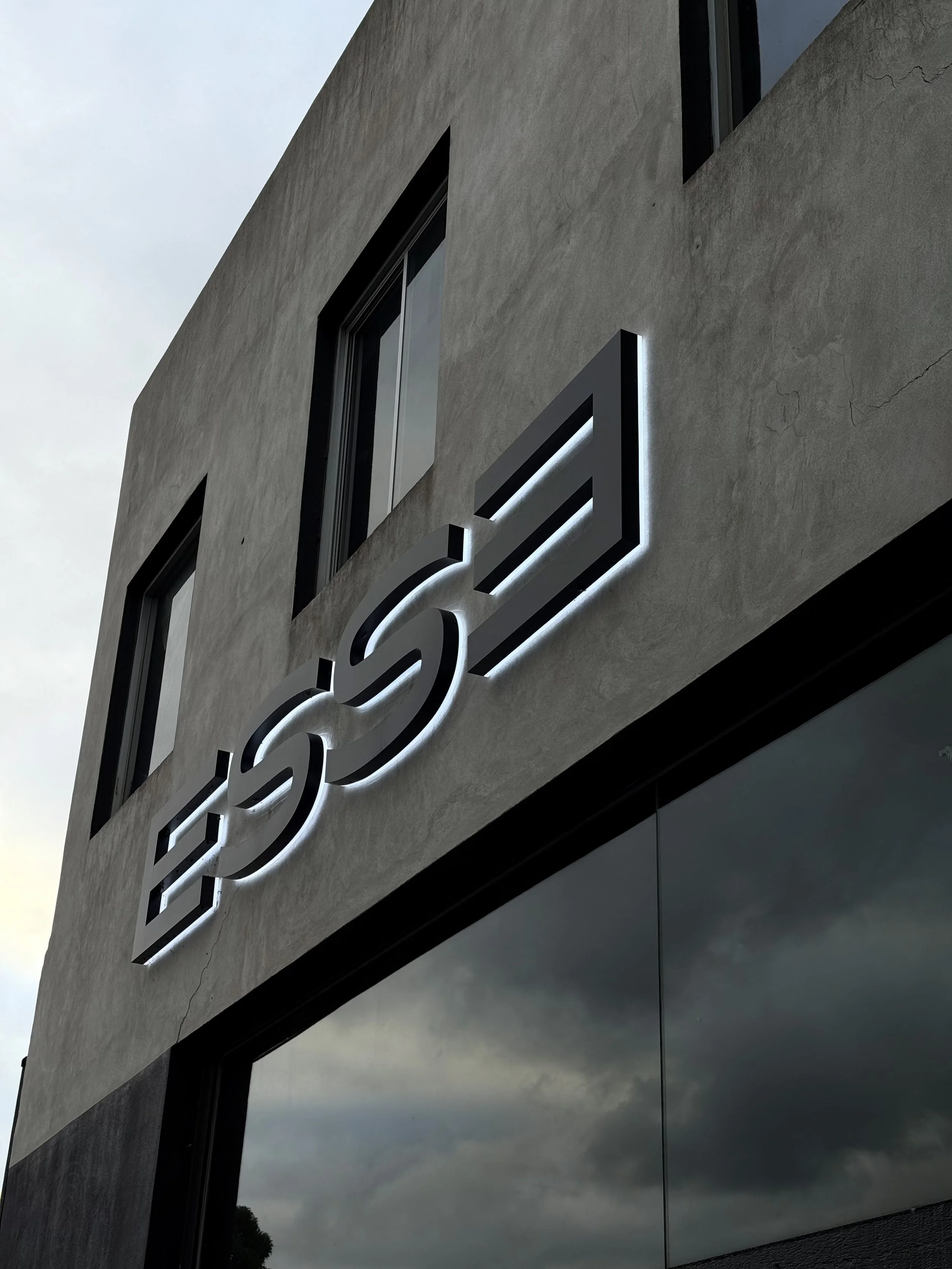 Close-up of a modern building's exterior with a sign displaying the 'ESSE' logo illuminated in white light against a gray concrete wall.