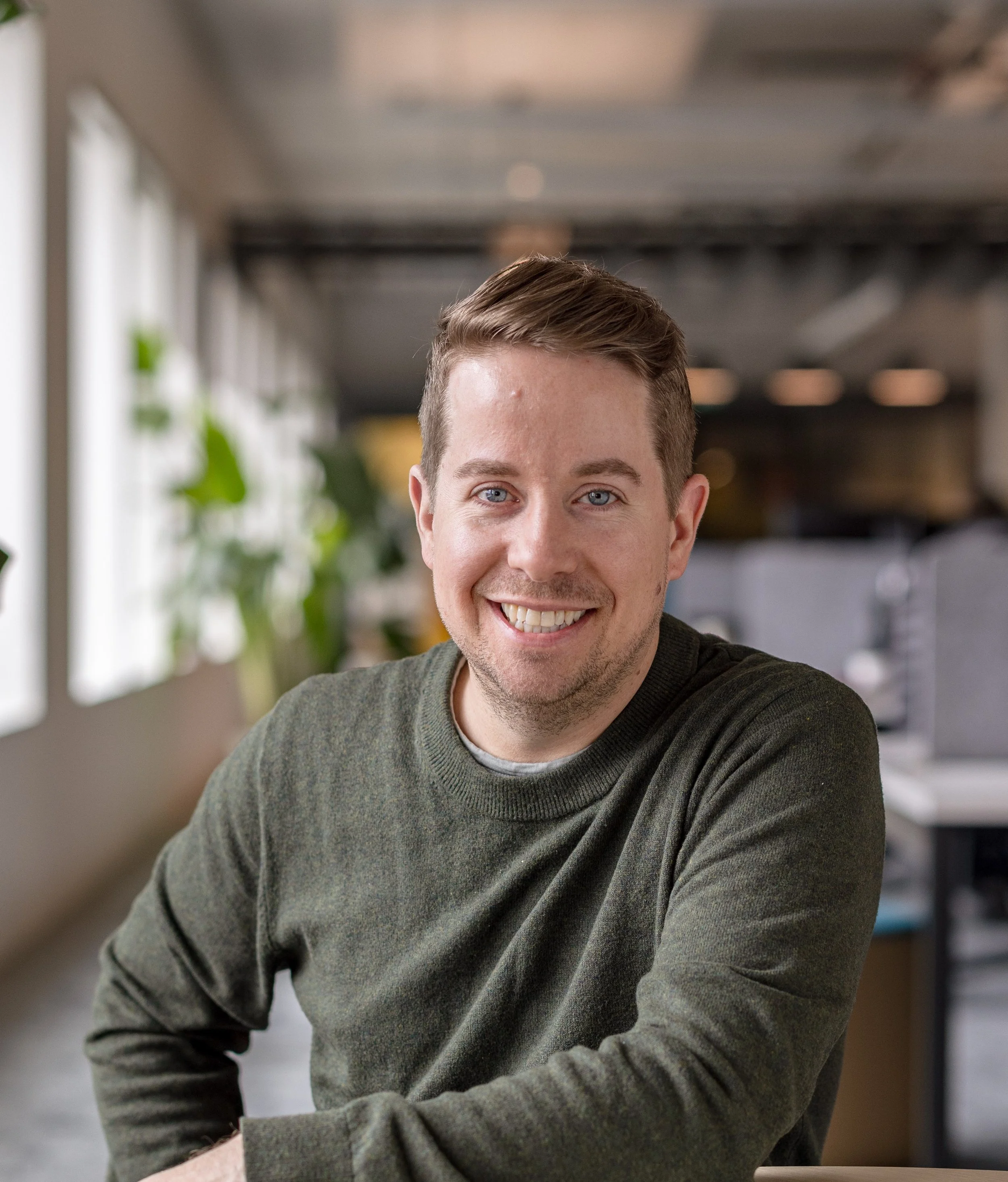 A smiling man, Chris Quinn, with short brown hair and blue eyes, wearing a dark green sweater, sitting in a modern office with large windows and plants in the background.