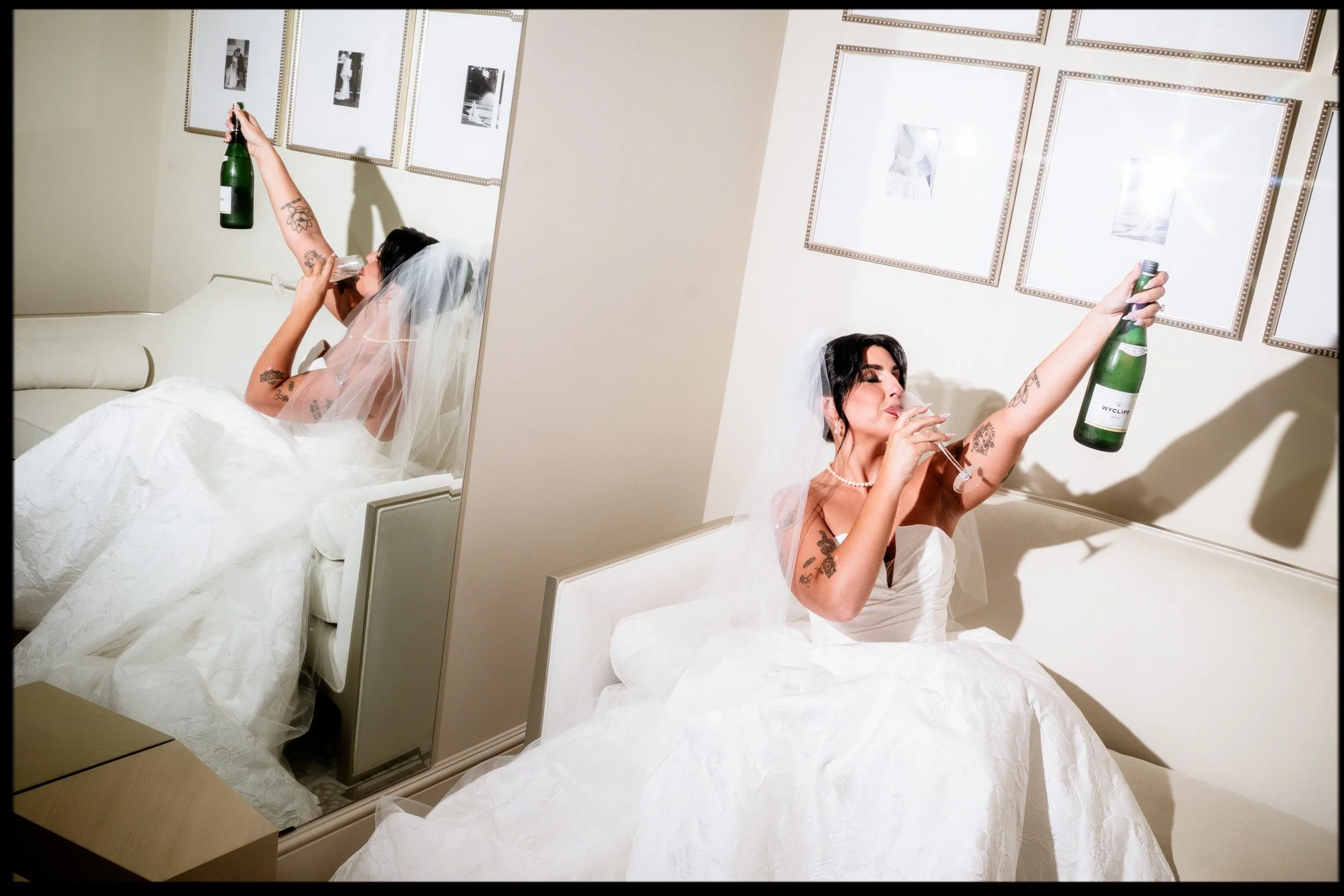 A bride in a wedding dress with a veil, seated on a bed, holding a champagne bottle in one hand and a champagne glass in the other, looking at the glass and smiling, with a mirror reflecting her image and framed artwork on the wall.