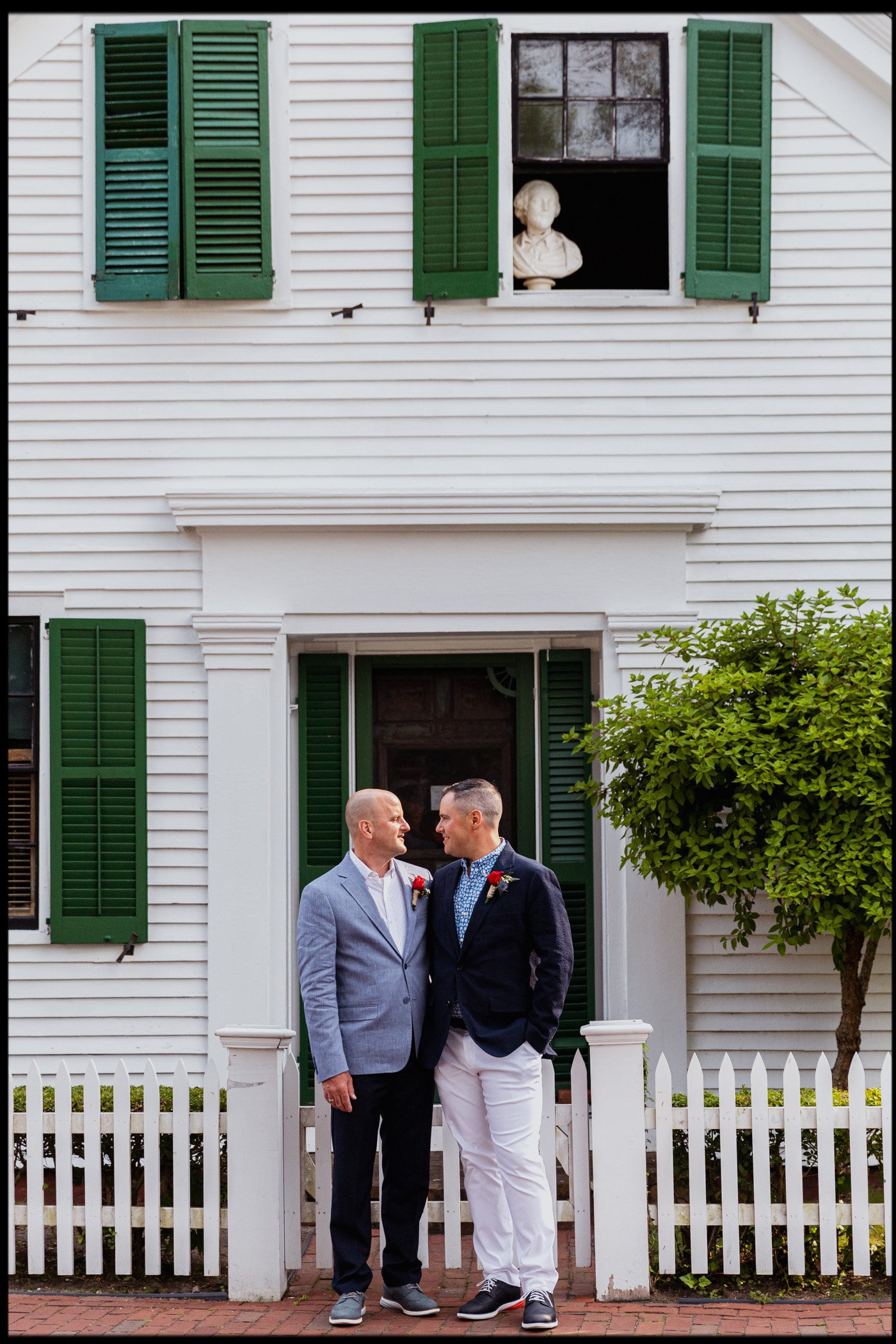 Two men standing in front of a white house with green shutters, dressed in suits, looking at each other. The house has a small white picket fence and a tree on the right side.