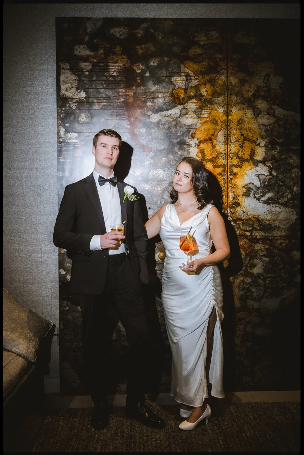A man and woman dressed in formal attire standing against an abstract textured background. The man is in a black tuxedo with a bow tie, holding a cocktail, while the woman is in a white gown holding an orange drink.