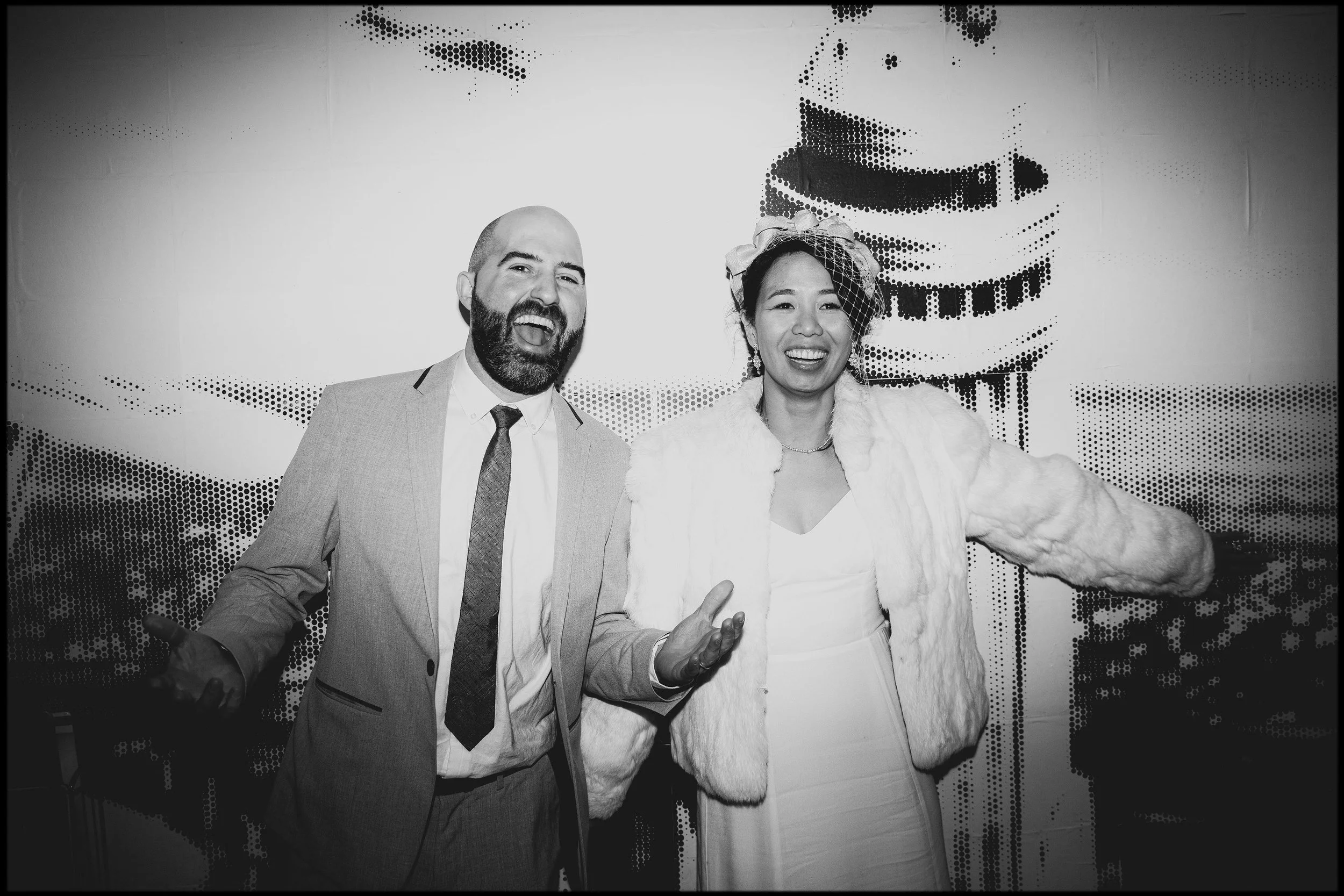 A man with a beard wearing a suit and tie, and a woman in a white dress with a fur coat and a fascinator, both smiling and posing in front of a graphic backdrop.