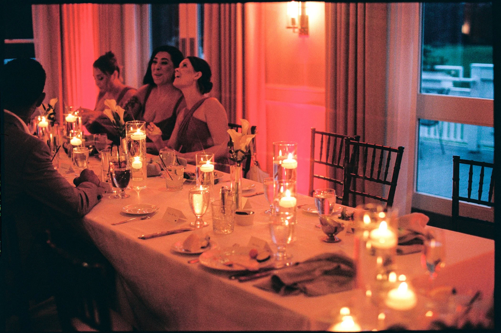 A group of women sitting at a formal dinner table in a warmly lit room with candles and elegant table settings, engaged in conversation and laughter.