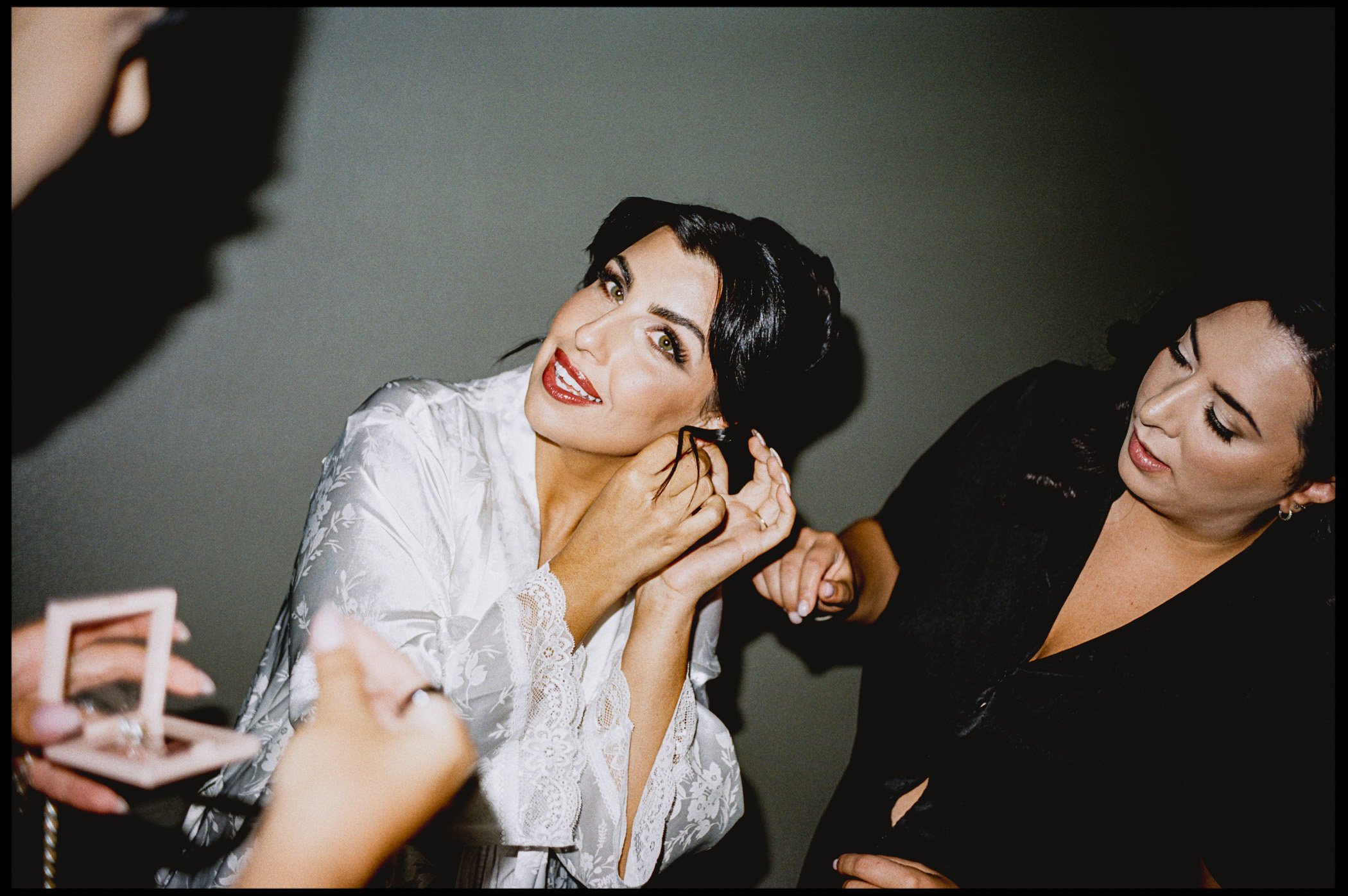 A woman with dark hair and bright lipstick, wearing a lace white dress, is smiling and adjusting her earring while another woman with dark hair and wearing a black dress helps her.