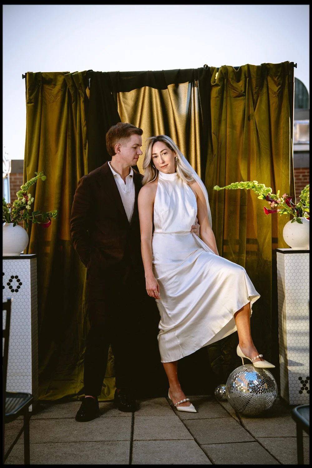 A man and woman posing together in front of a gold curtain backdrop, with decorative plants on white stands, on a tiled rooftop or terrace.