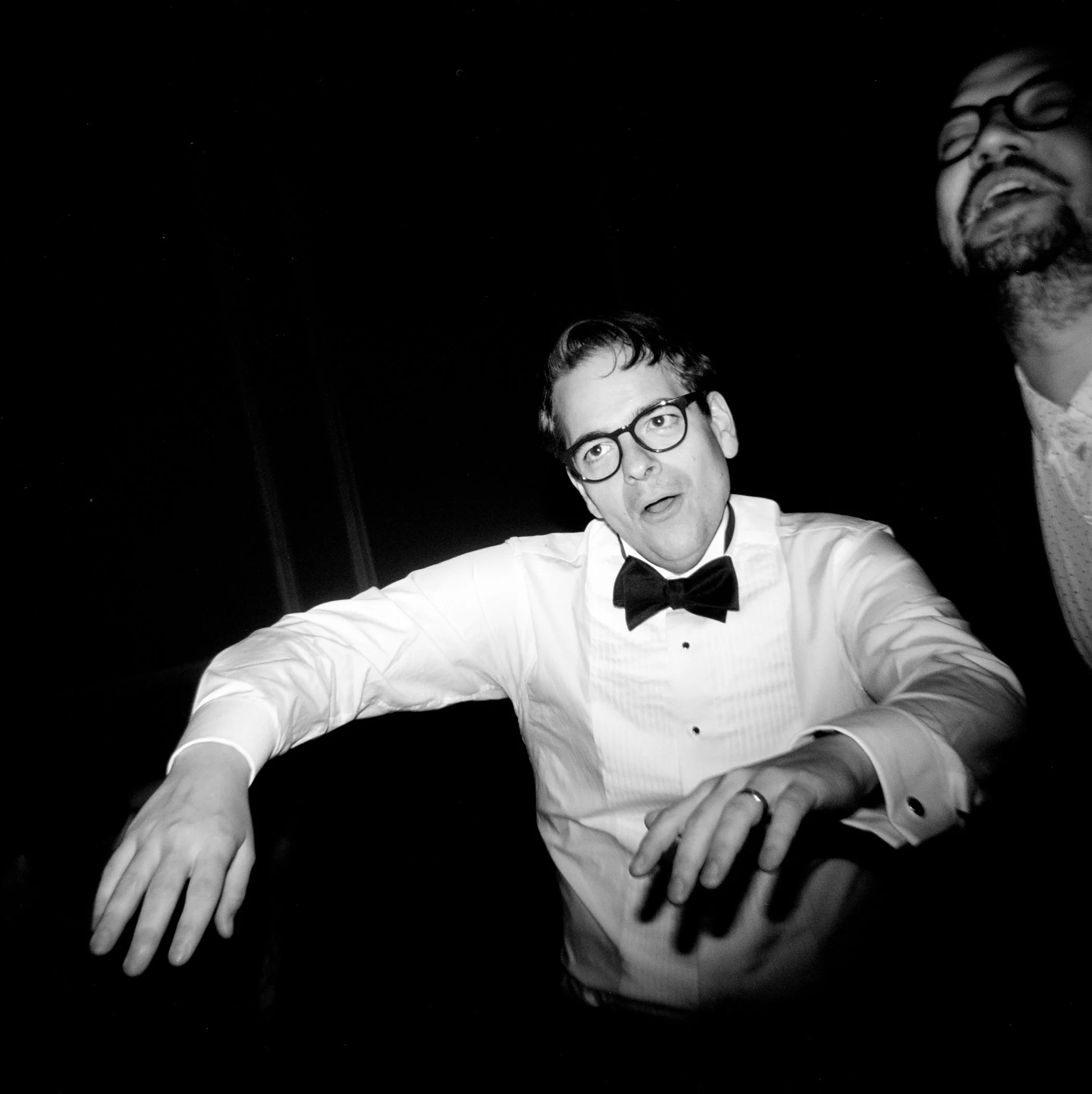 Black-and-white photo of two men in tuxedos. One man is looking directly into the camera and the other is smiling, both behind multiple outstretched hands.