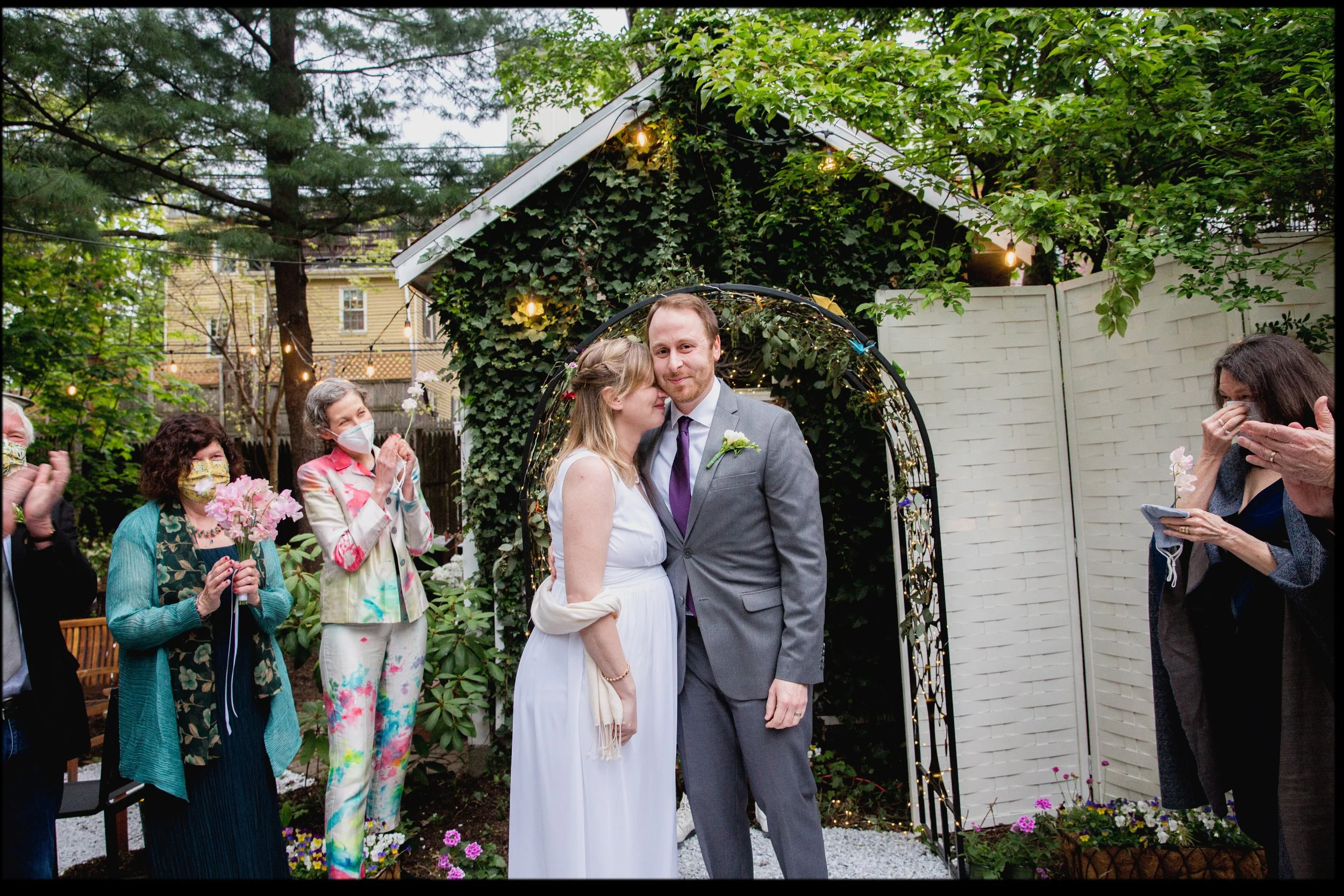 A couple gets married outdoors surrounded by friends and family. The bride wears a white dress, and the groom wears a gray suit. Celebrants are clapping and smiling, with some wearing face masks. There are decorative lights and green foliage in the background.