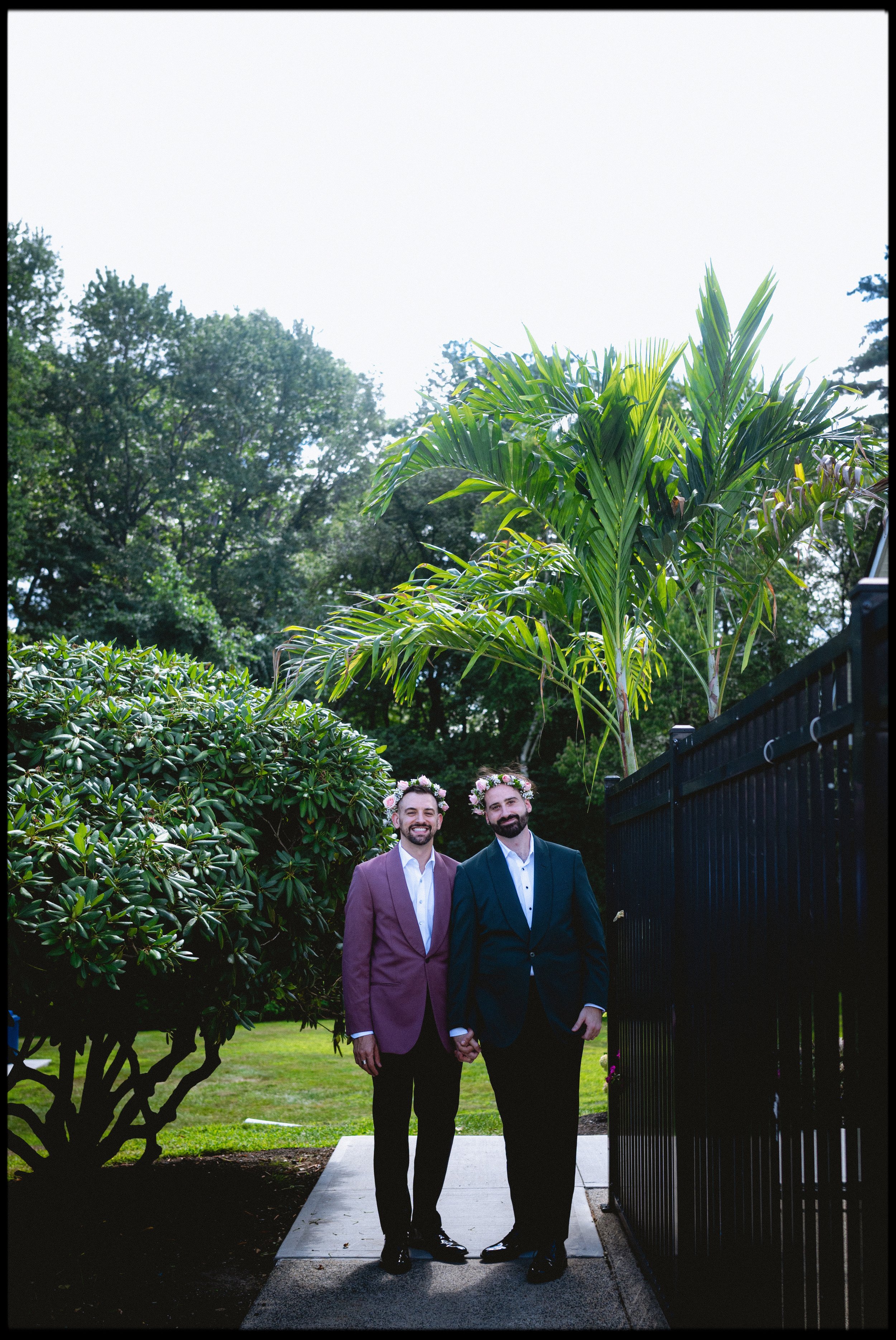 Two men in suits holding hands and wearing floral crowns walking on a pathway in a garden, with trees and bushes in the background.